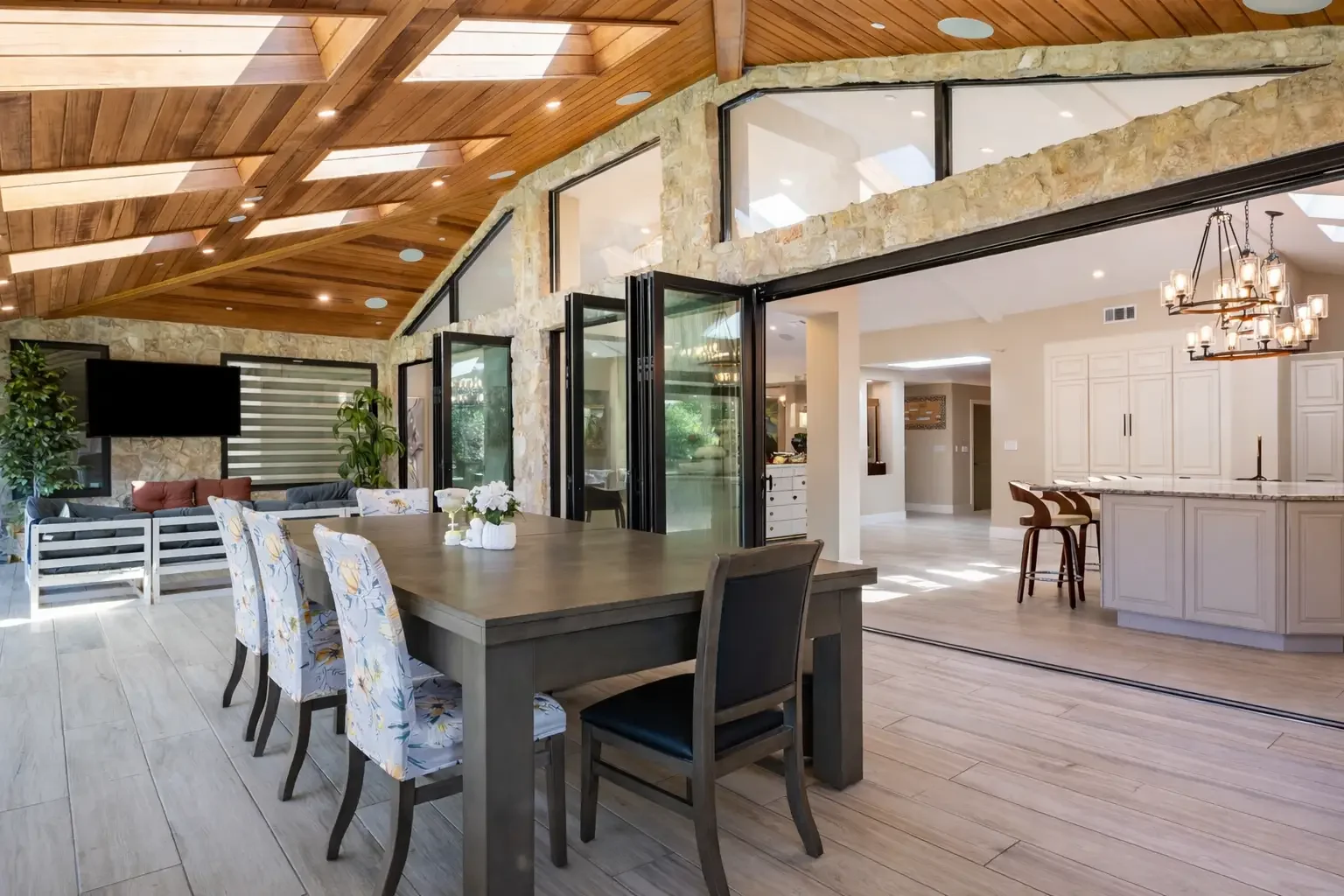 Open-concept dining and living room with wooden ceiling, stone wall, large windows, and sliding glass doors leading to a bright kitchen with white cabinetry and a chandelier.