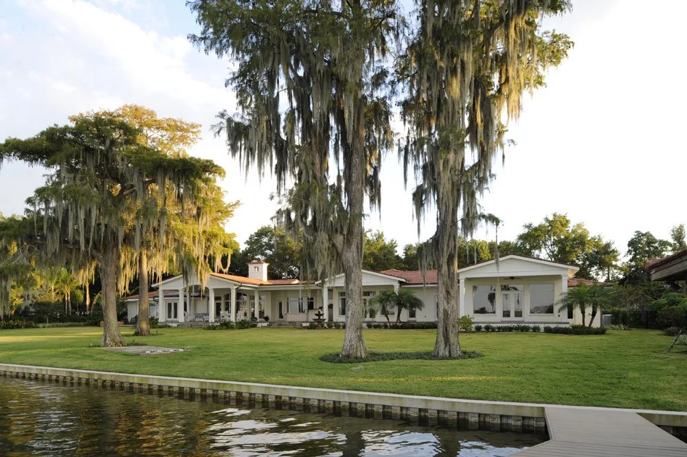 A large, white house with expansive windows, surrounded by lush green grass and trees draped with Spanish moss, next to a body of water with a small dock in the foreground.