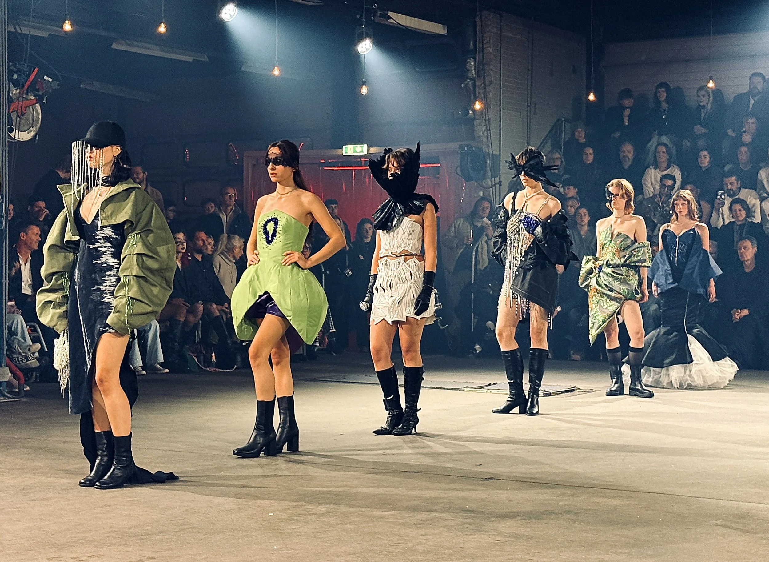 Models walking on a runway during a fashion show, wearing avant-garde and modern clothing, with an audience seated on both sides.