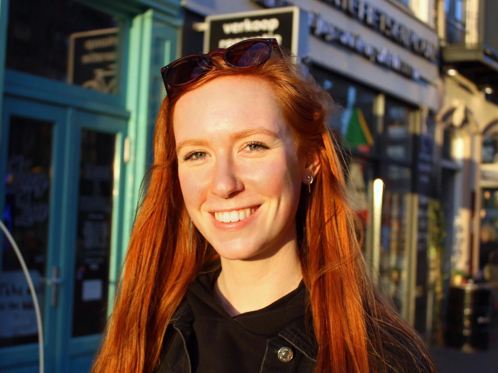 A young woman with red hair smiling outdoors, with sunglasses on her head and wearing a black jacket, in an urban setting during sunset.