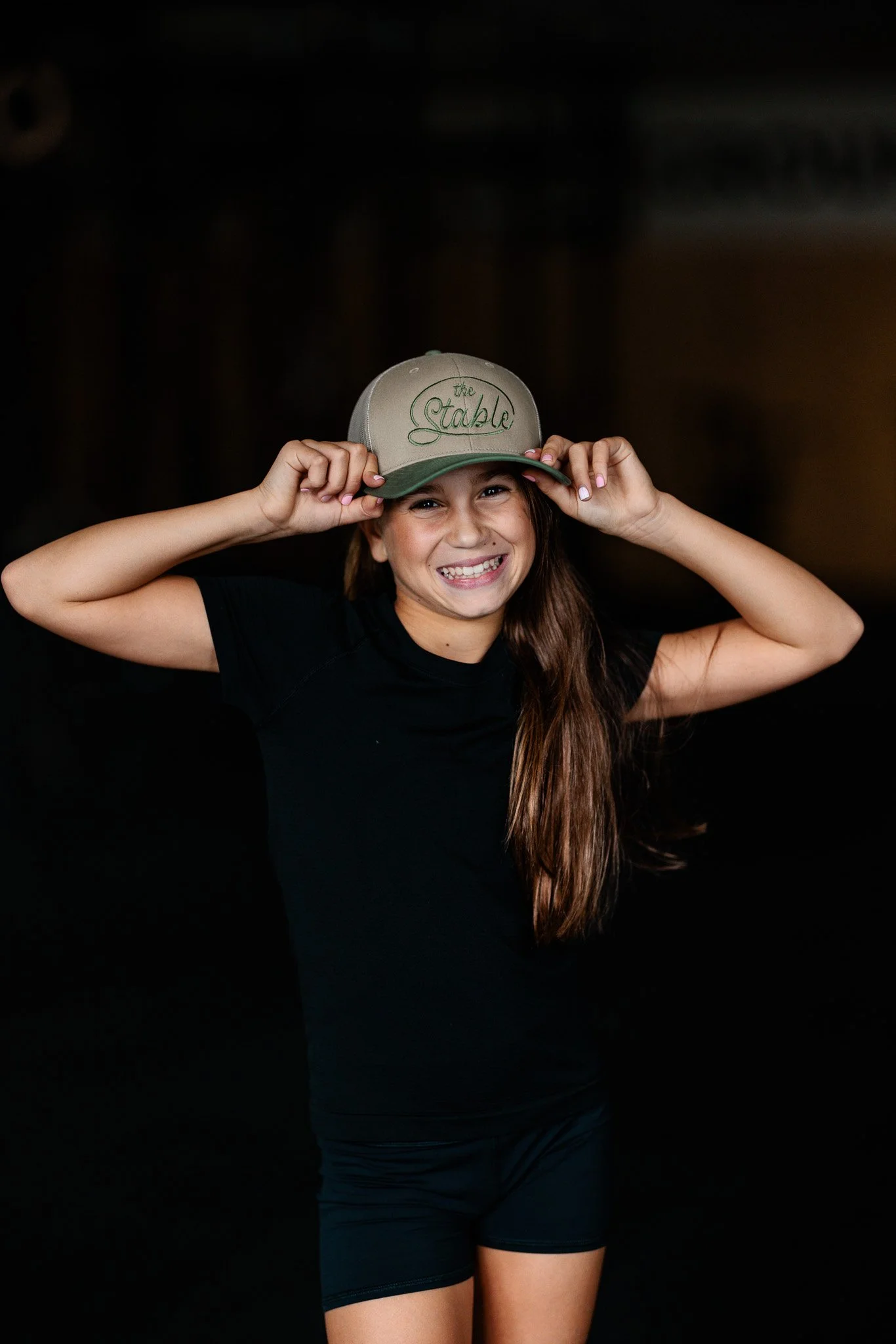 A young girl smiling at the gym, wearing a hat that says 'the STABLE strength & conditioning,' with black clothes on in a fitness center.