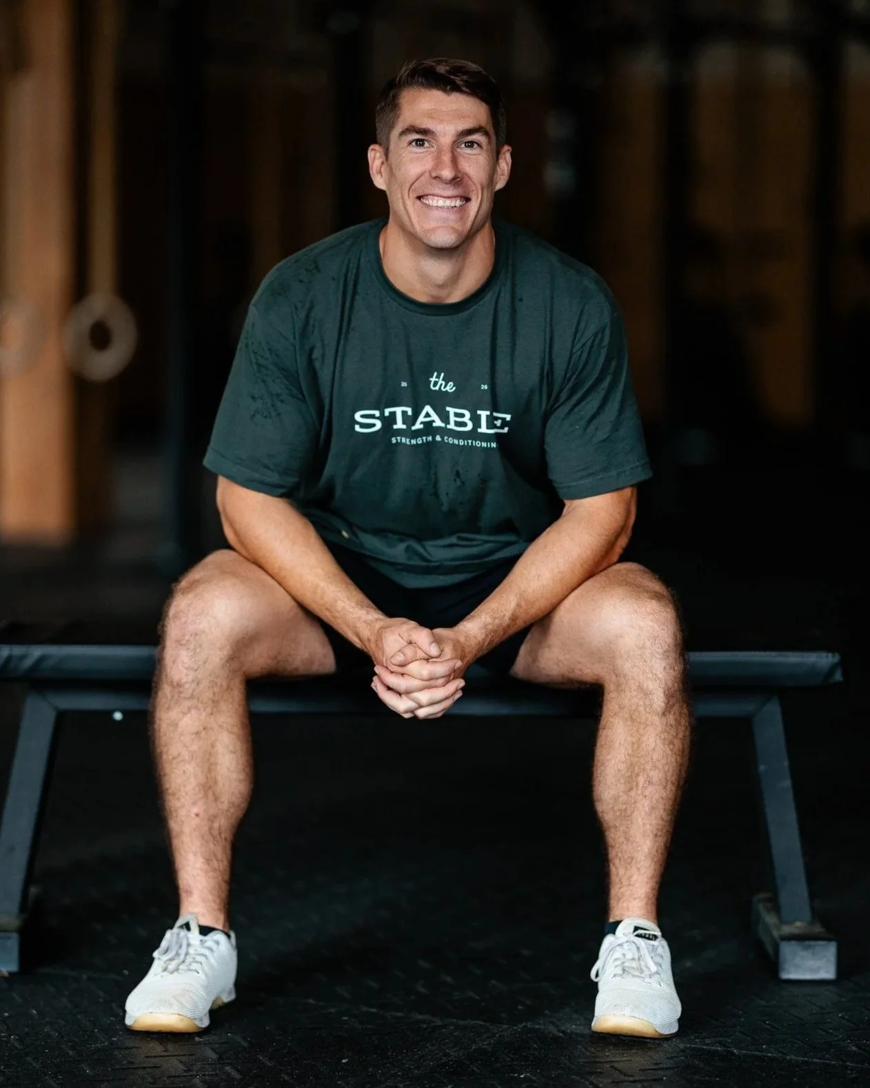 A man sitting on a workout bench in a gym wearing a gray tshirt that say "The Stable Strength & Conditioning"