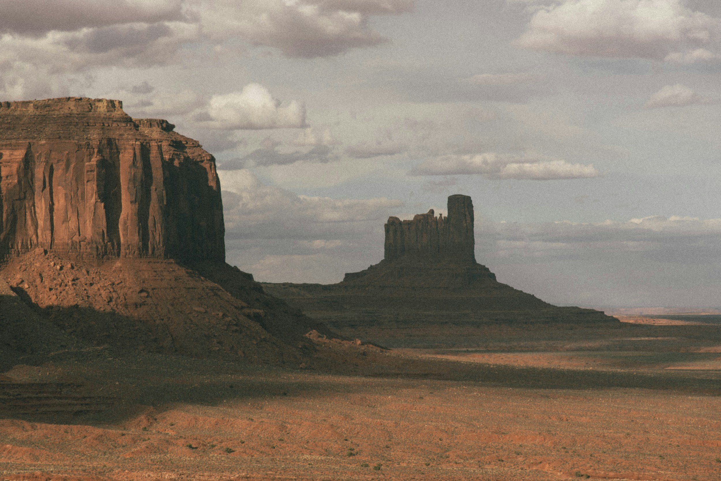 A desert landscape with large rock formations and mesas under a cloudy sky.