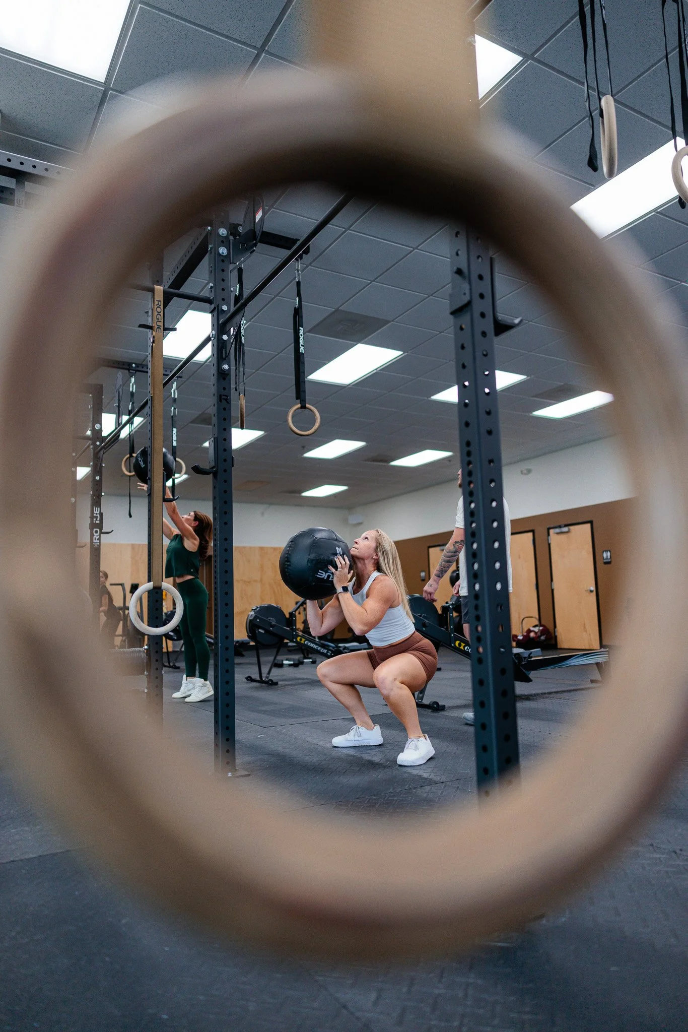 A woman with blonde hair doing a wall ball squat in a fitness center with a Rogue rig and hanging rings.