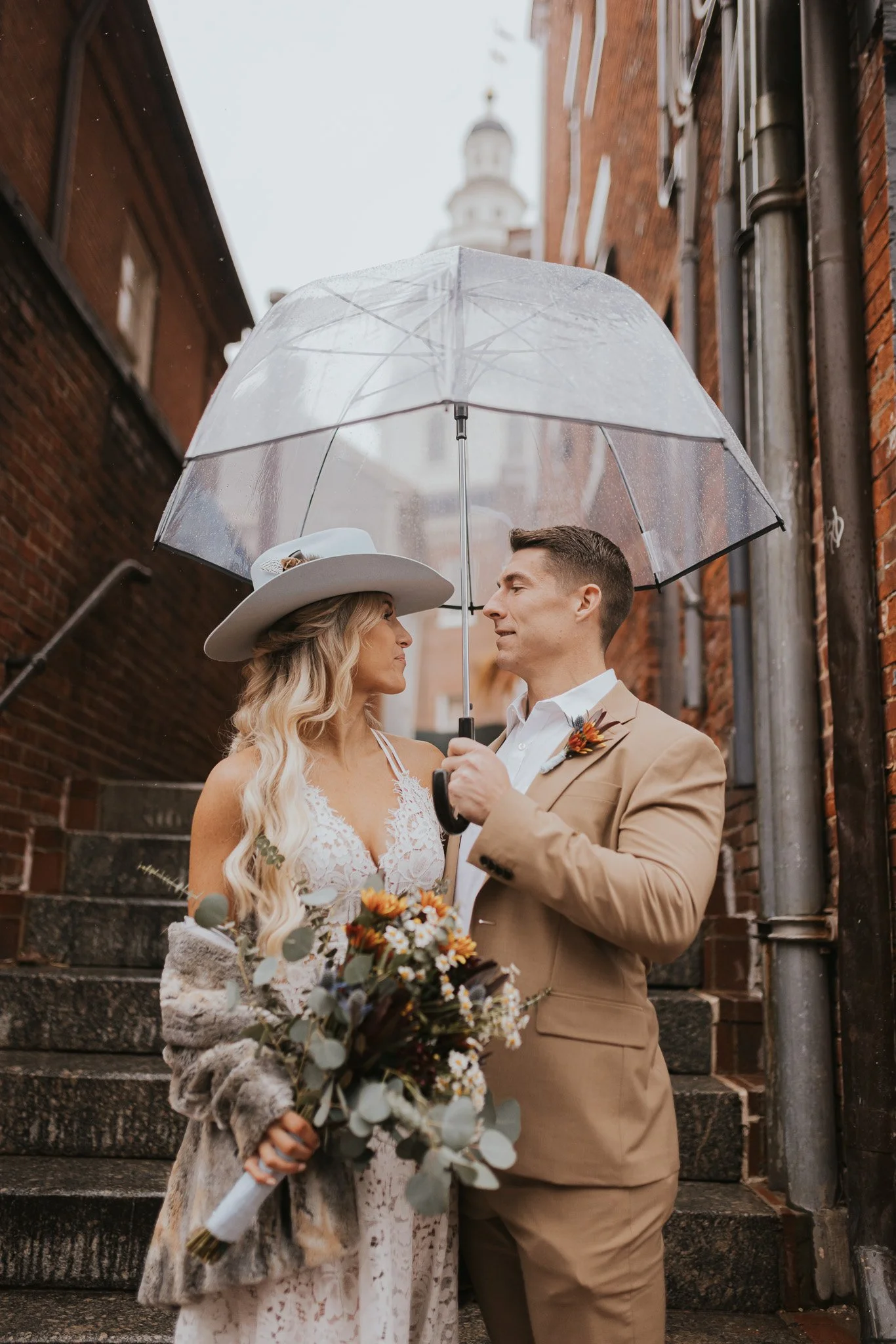 A bride and groom stand close under a clear umbrella on a rainy day in an alley with brick buildings and blurred church steeple in the background. The bride wears a white lace dress and a large hat, holding a bouquet of flowers. The groom wears a tan suit with a boutonniere, holding the umbrella over both.