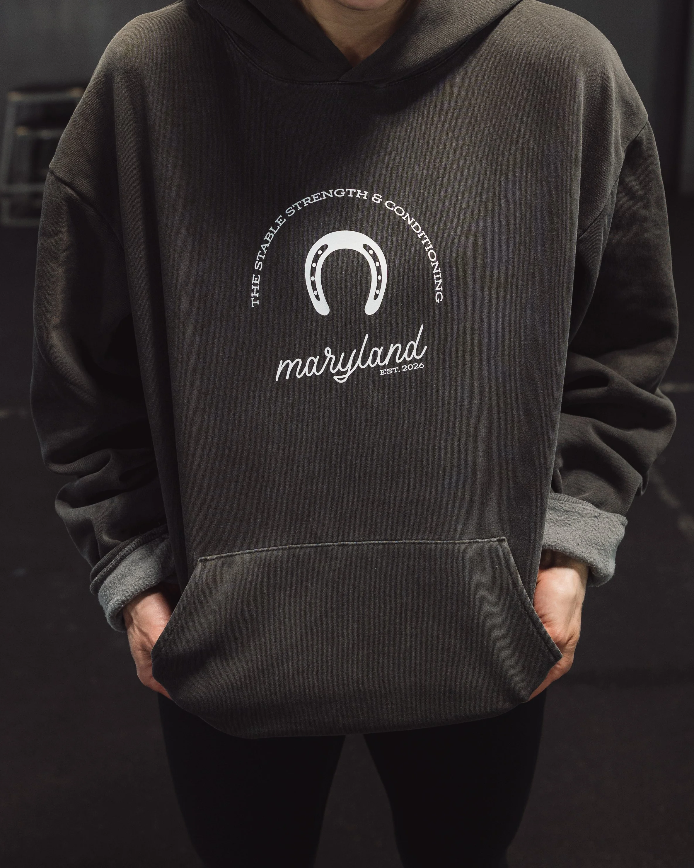 A person wearing a gray hoodie with the words 'The Stable Strength & Conditioning Maryland' and a horseshoe logo on the front, standing with hands in front pockets.