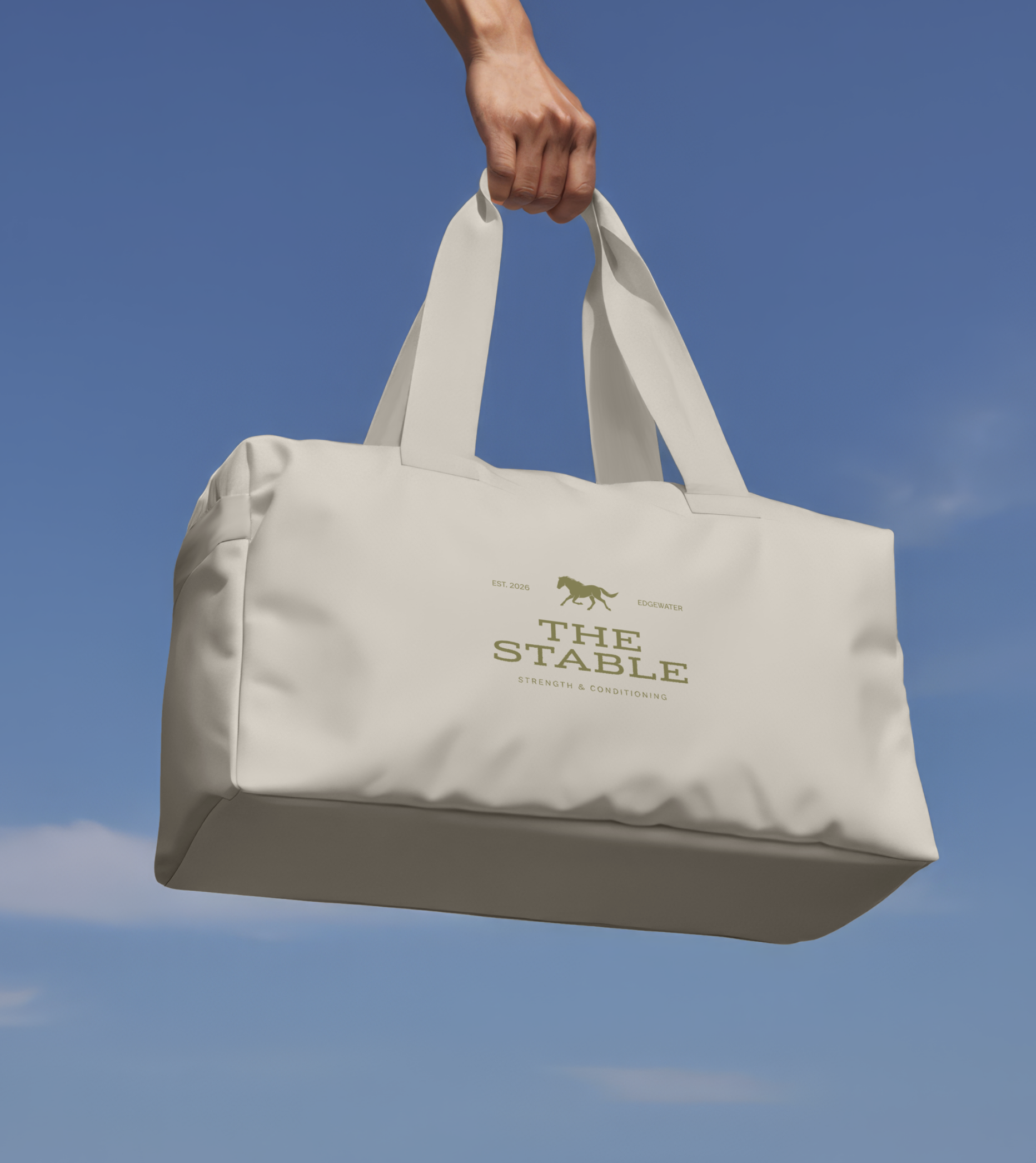 A person's hand holding a white tote bag with the text 'The Stable Strength & Conditioning,' a horse silhouette, and the words 'Est. 2026' printed on it, against a blue sky background.
