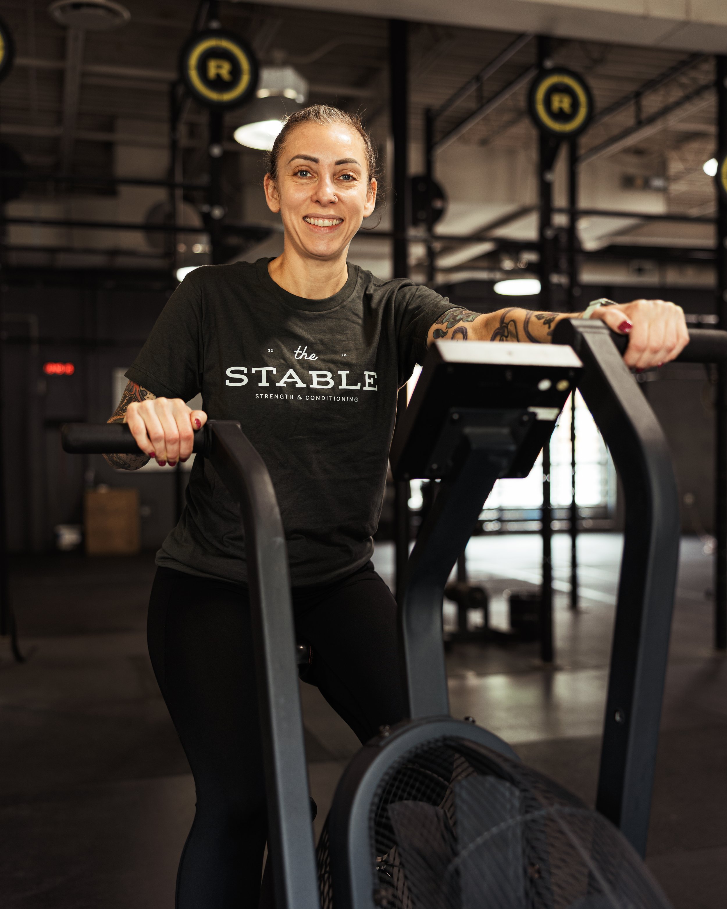 A woman smiling at the gym, wearing a black t-shirt that says 'the STABLE strength & conditioning,' with tattoos on her right arm, standing behind a rowing machine in a fitness center.