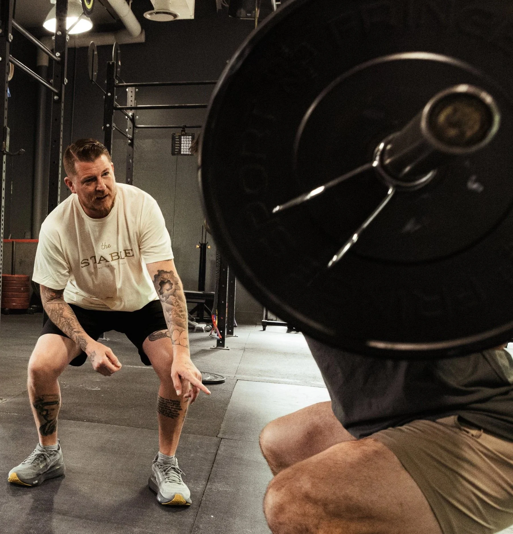 A man with tattoos on his arms and legs is in a gym, wearing a white T-shirt, black shorts, and sneakers. He appears to be in a workout or training session. Large black fitness equipment or weights are visible in the foreground, with gym walls and equipment in the background.