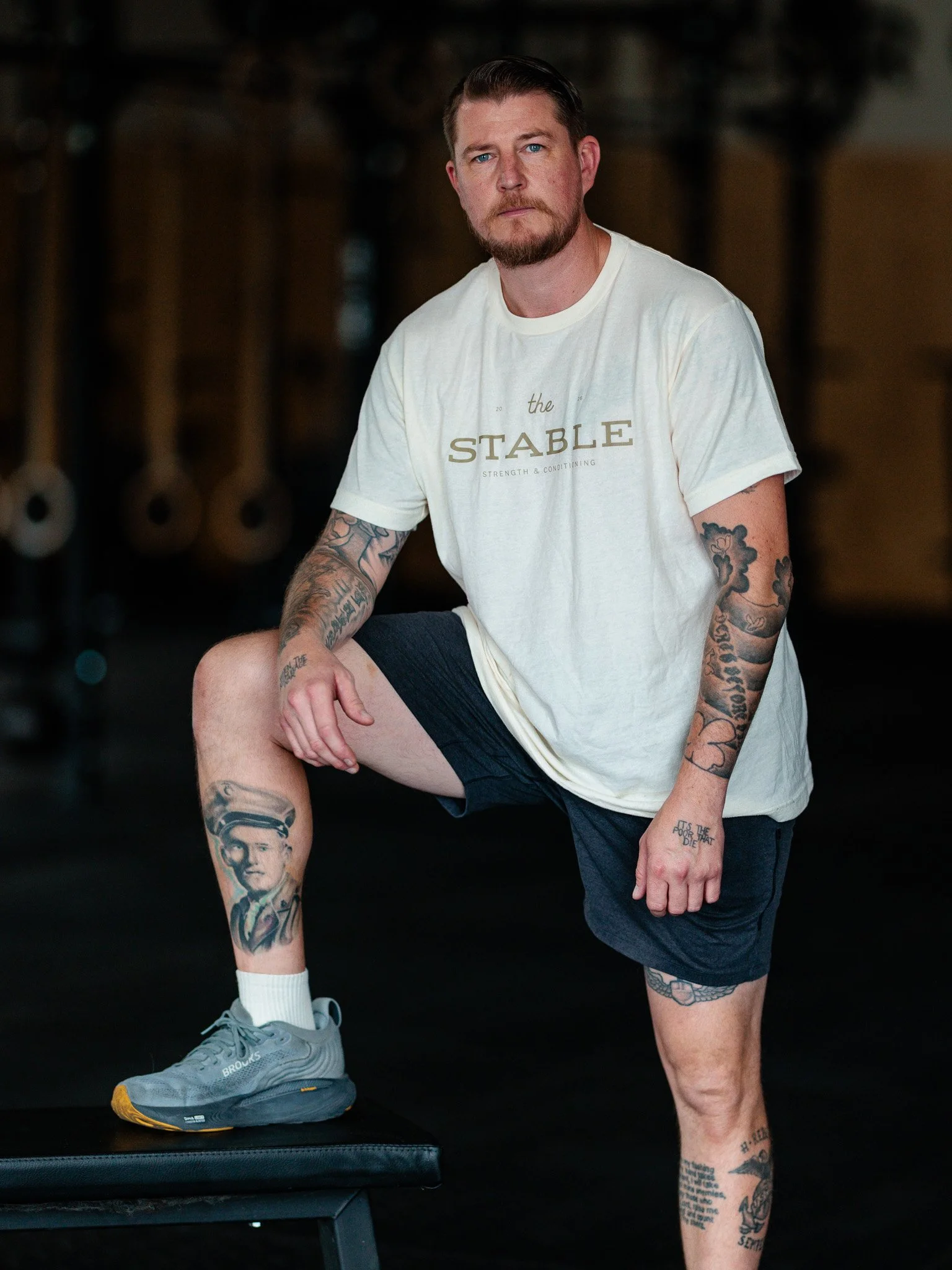A man with tattoos standing with one leg up on a workout bench in a cream tshirt that say "The Stable Strength & Conditioning"