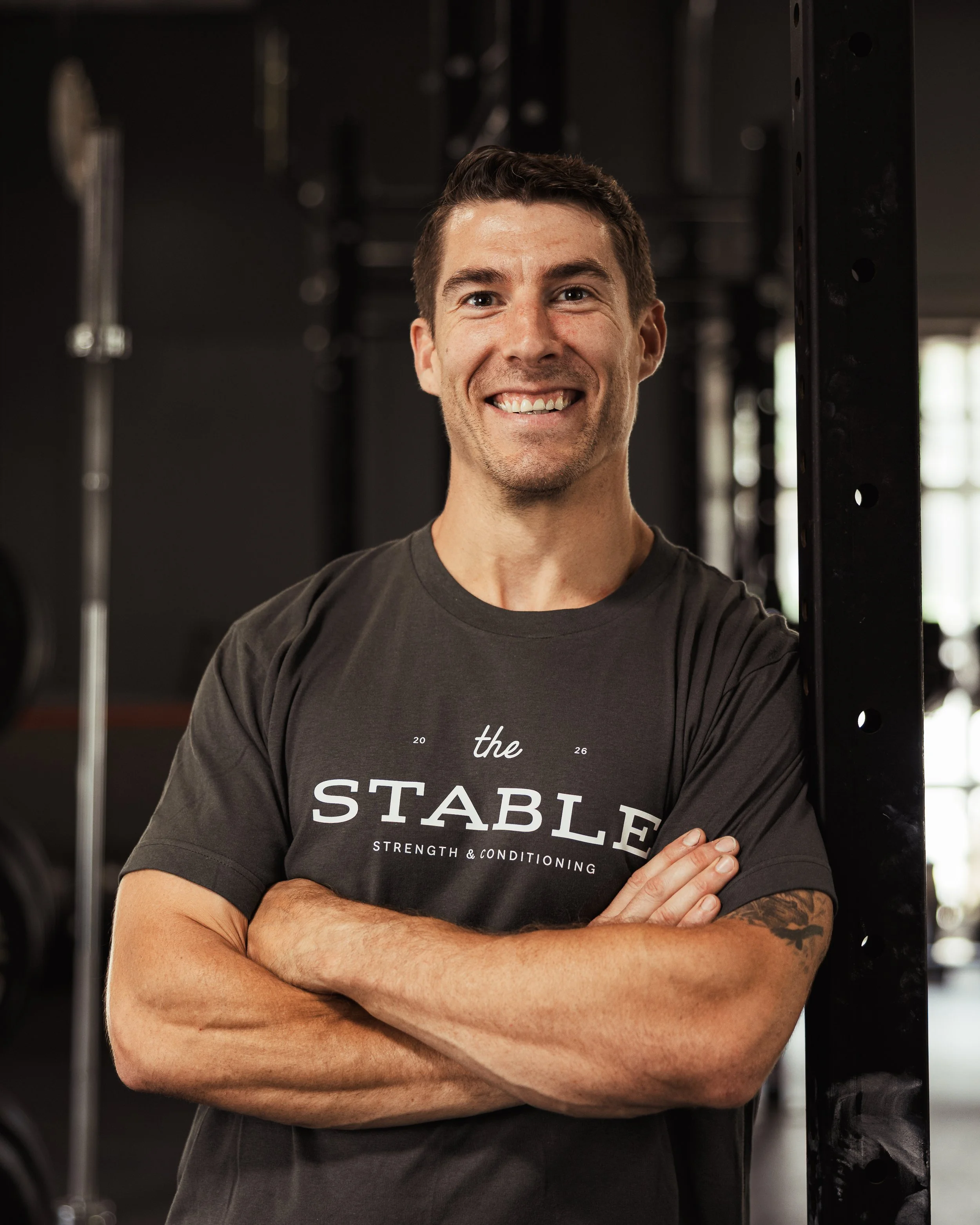 A smiling man with crossed arms standing in a gym, wearing a black T-shirt with the words "the STARTLE" visible, surrounded by gym equipment.