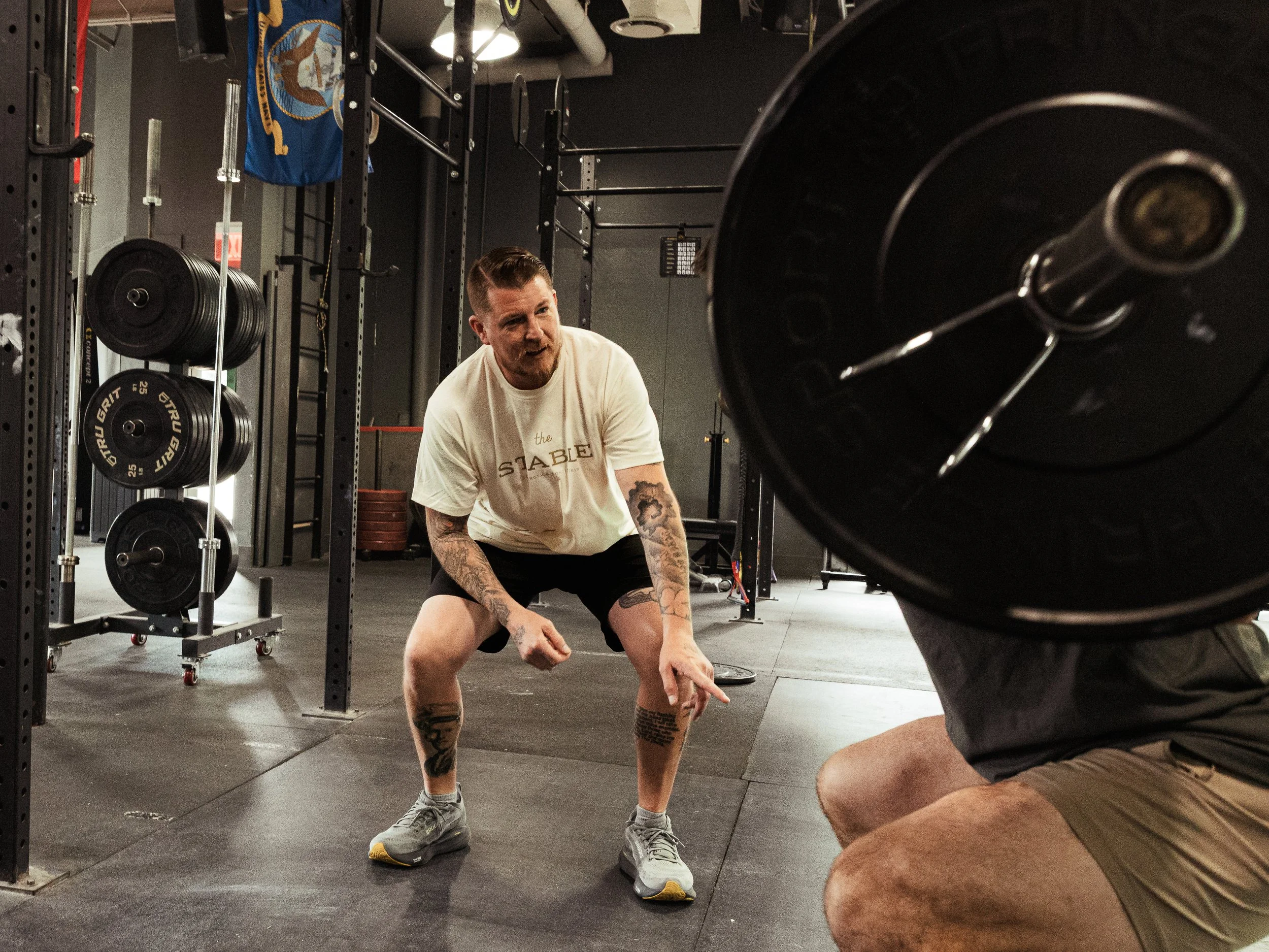 A man with tattoos wearing a white T-shirt and shorts is in a gym, bending down to pick up a weight. There are weight plates on a rack in the background, and part of another person is visible in the foreground, wearing brown shorts and a gray shirt.