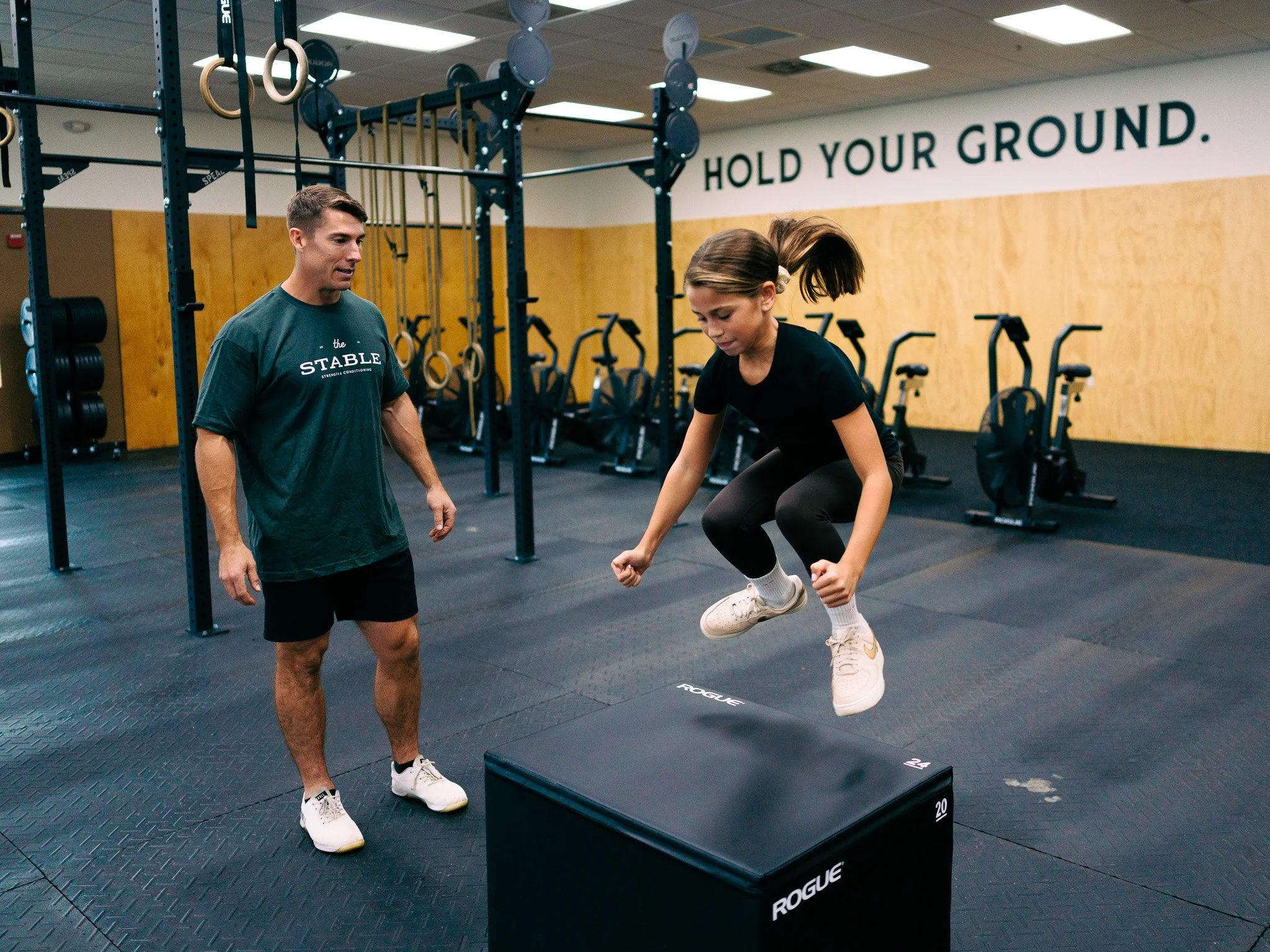 A man in a gray tshirt that says "The Stable Strength & Conditioning" instructing a youth girl on how to perform a box jump in a gym.