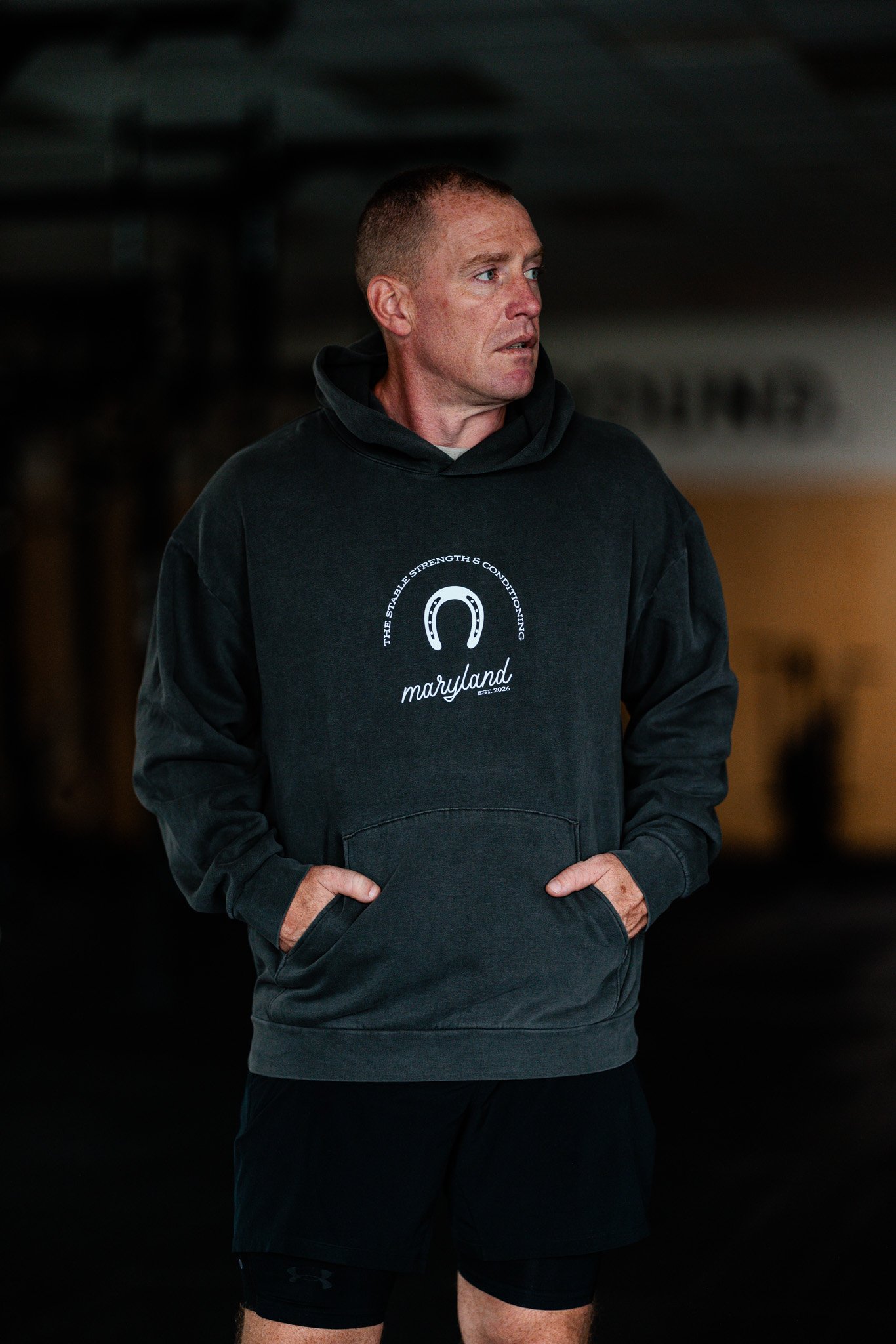 A man standing in a dark gym wearing a charcoal hooded sweatshirt that has a horseshoe on it and says "The Stable Strength & Conditioning, Marlyand Est. 2026".