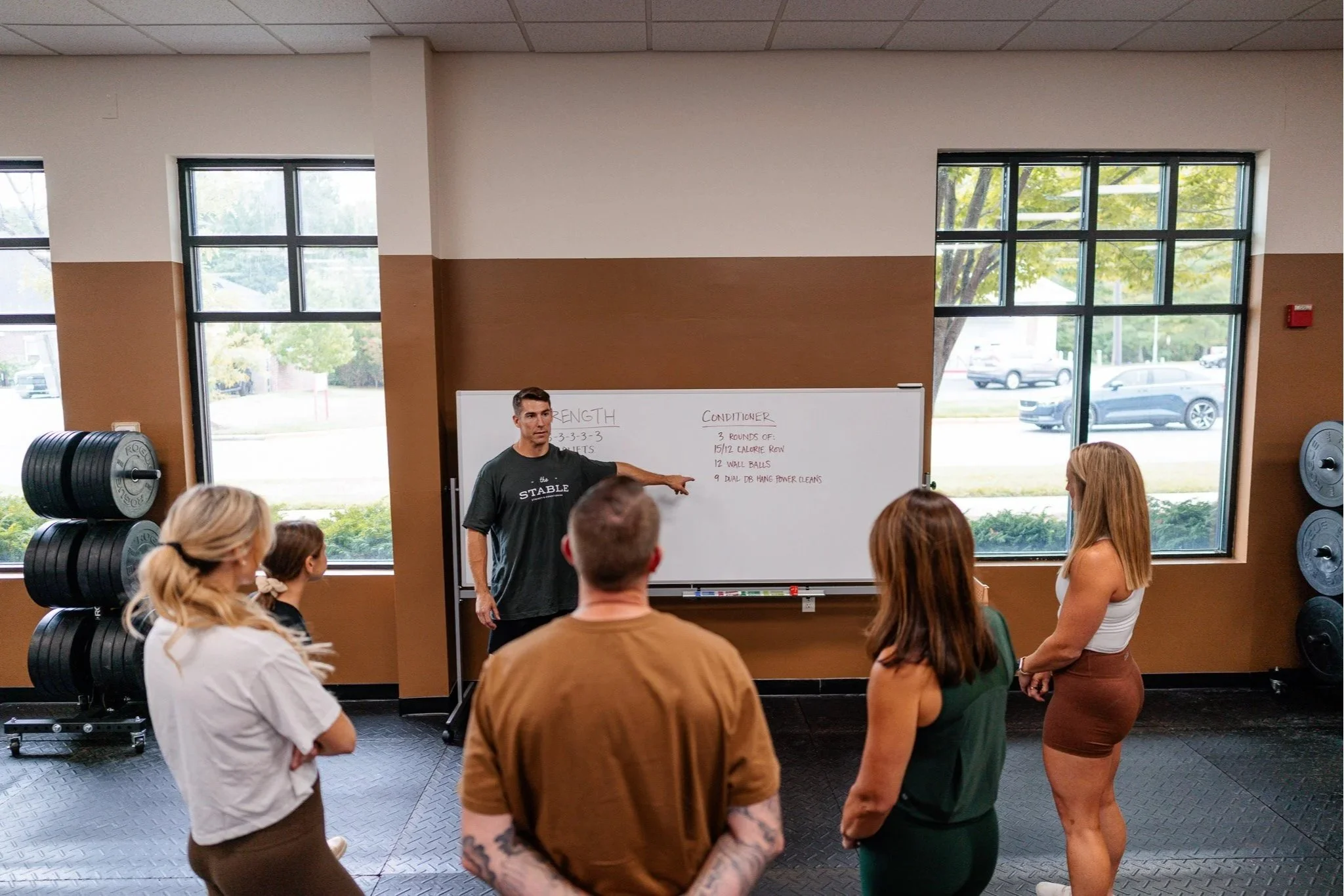 A man in a gray "The Stable" tshirt instructing members of a group fitness class in front of a whiteboard in a gym.
