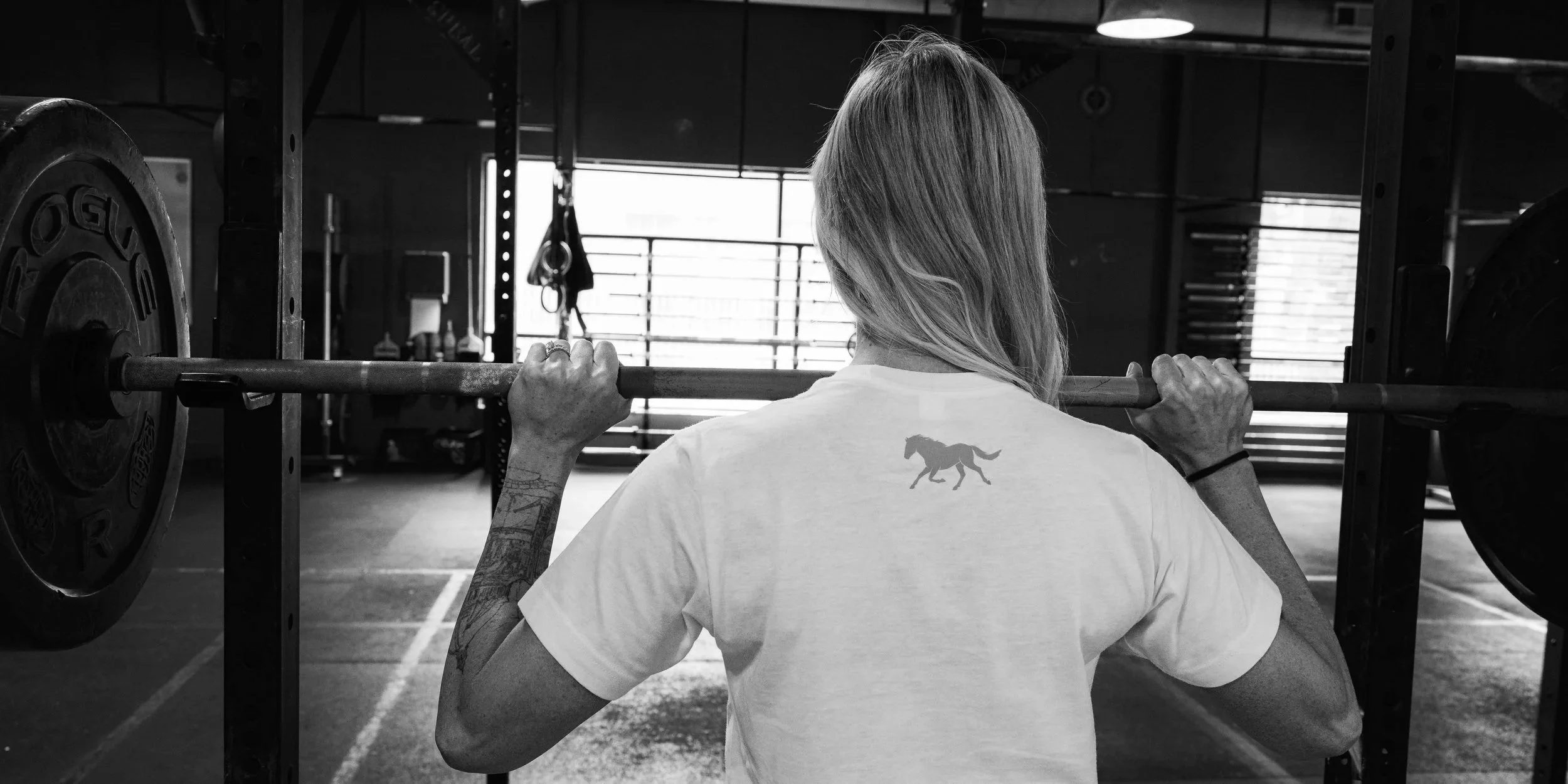 A woman with long hair, wearing a T-shirt with a horse silhouette, is standing in a gym lifting a barbell on her shoulders, with weight plates on each end.