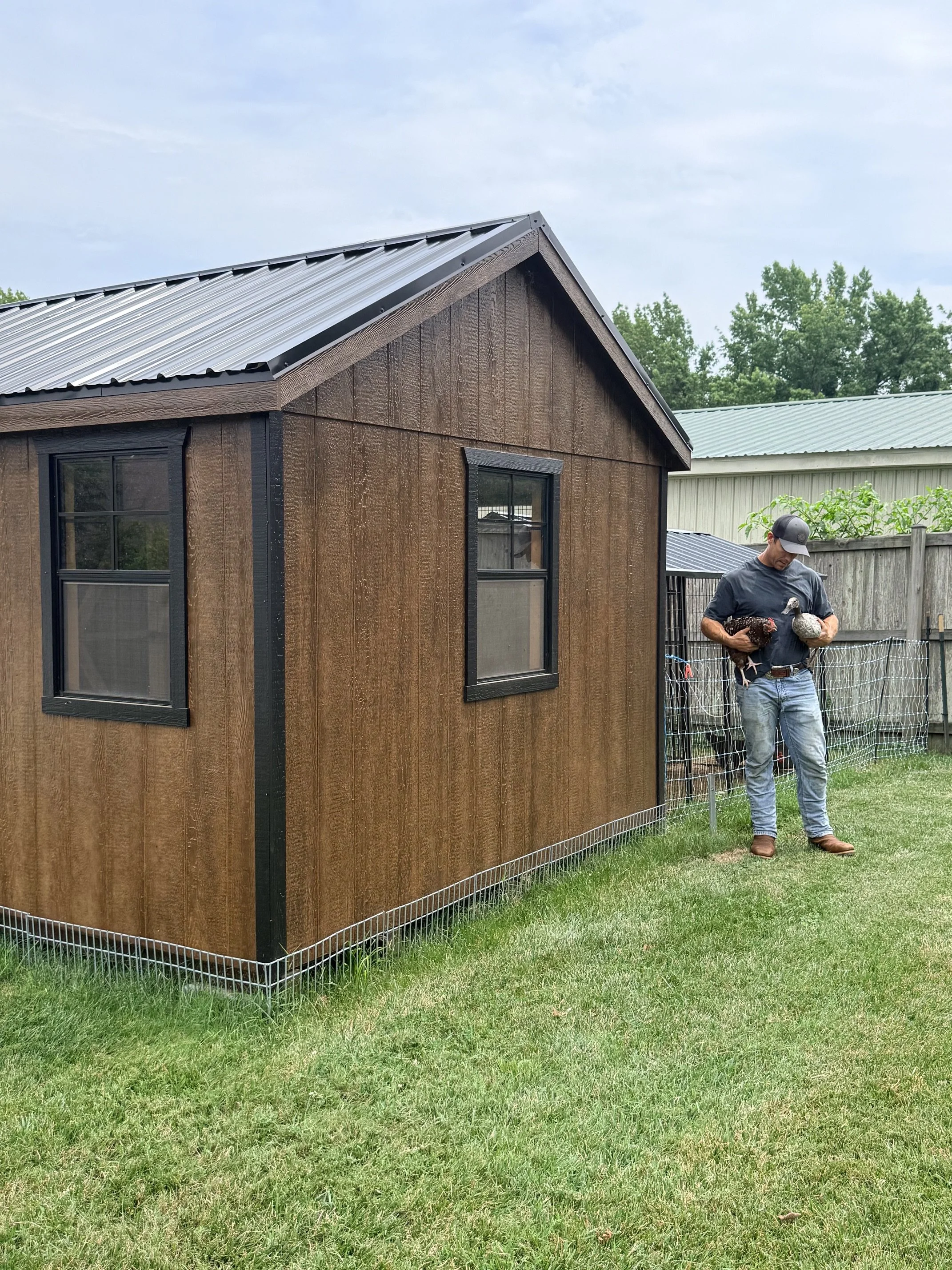 A man holding two chickens, one black and the other white, standing on a green yard near a wooden coop with black-trimmed windows and a metal roof, under a partly cloudy sky.