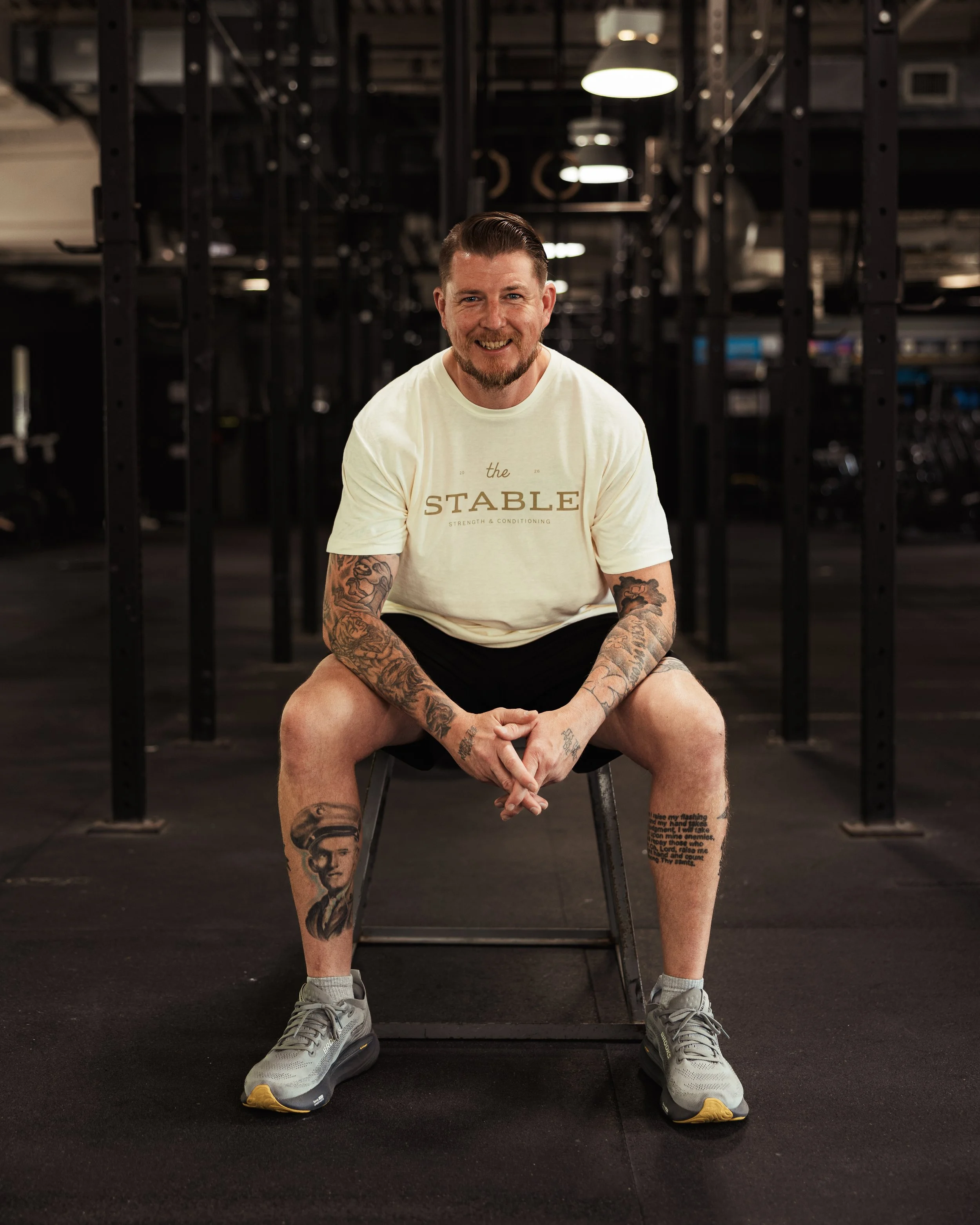 A man sitting on a chair in a gym, smiling at the camera. He has tattoos on his arms and legs, is wearing a white t-shirt with "the STABLE" printed on it, black shorts, and sneakers. The background shows gym equipment and black metal frames.