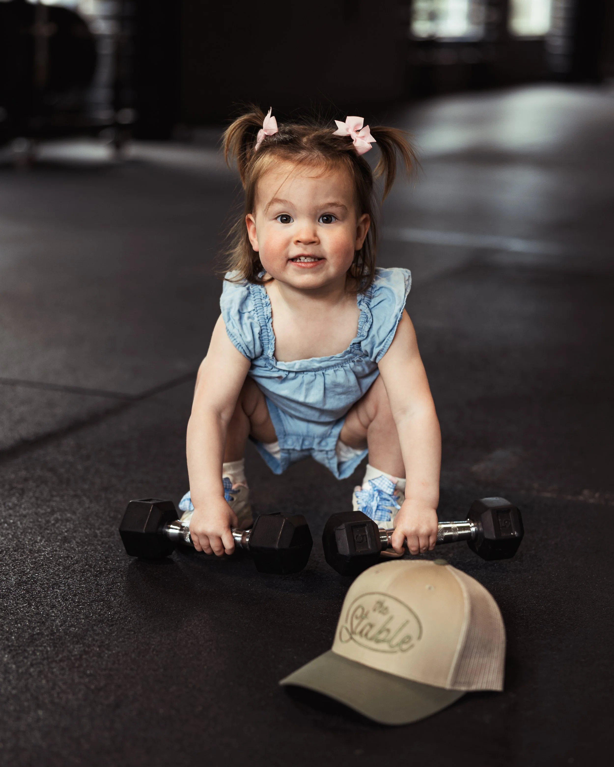 Young girl with pigtails and pink bows in a gym or fitness studio, holding black dumbbells on a black rubber floor, with a blurry background. A wooden rolling pin is partially visible in the foreground.