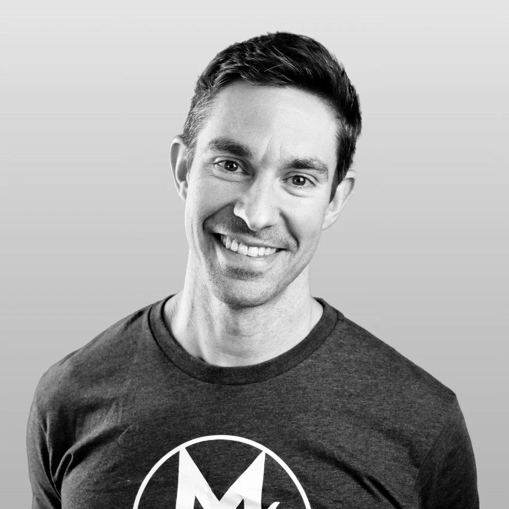Black-and-white photo of smiling man with short dark hair wearing a dark T-shirt with a circular logo.