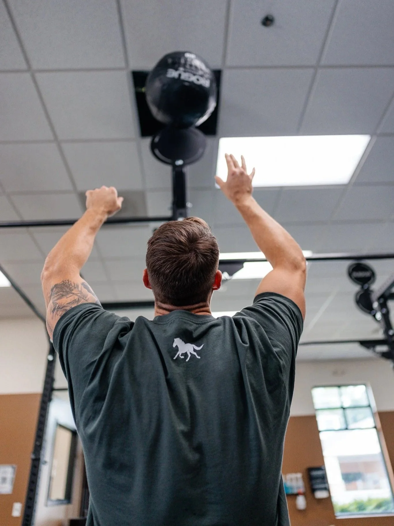 A man in a gray tshirt with a horse logo on the back of the neck doing a wall ball to a target on a Rogue rig.