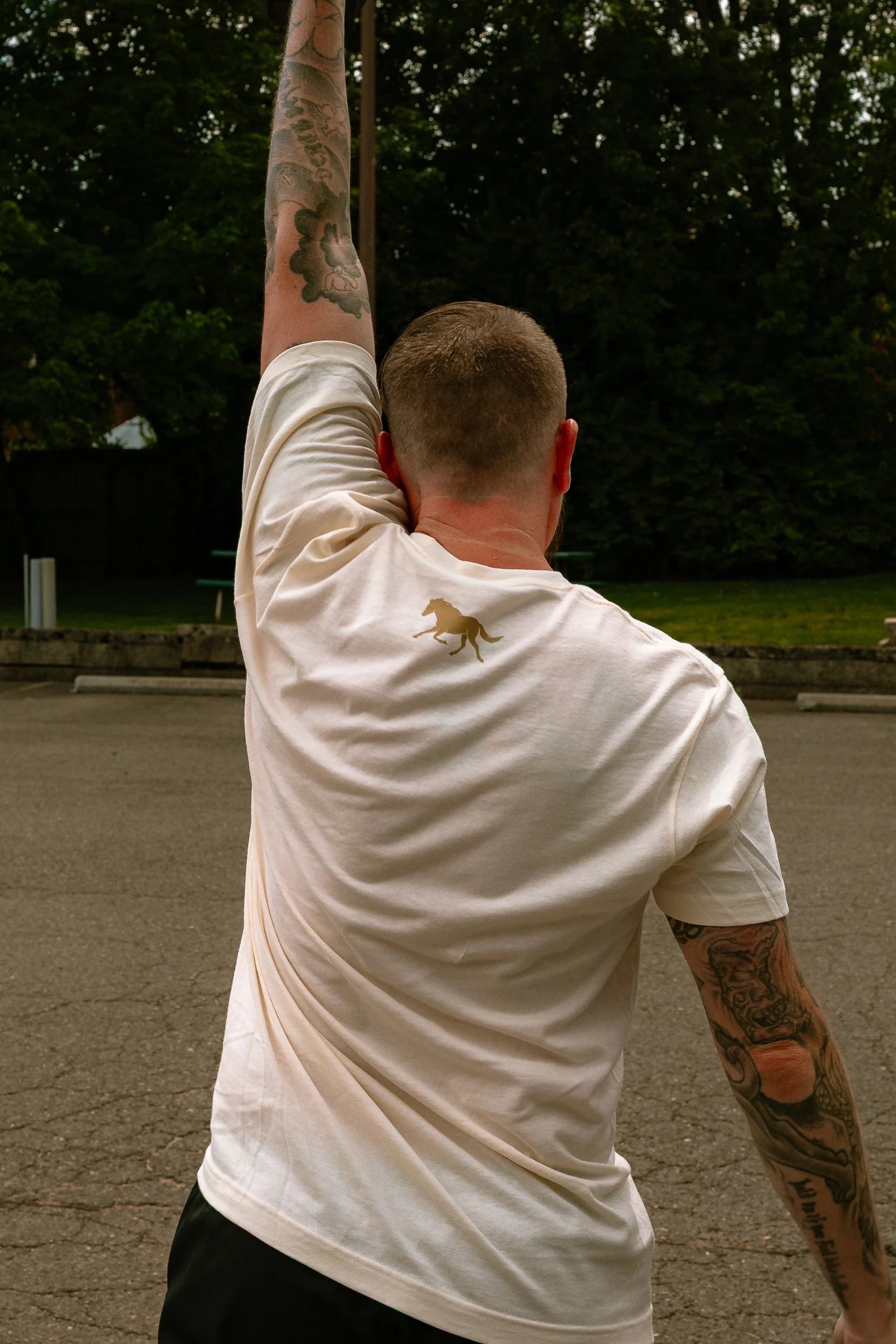 Rear view of a man with short hair and tattoos on his arm, wearing a white T-shirt with a horse logo, raising his arm in a park with trees in the background.