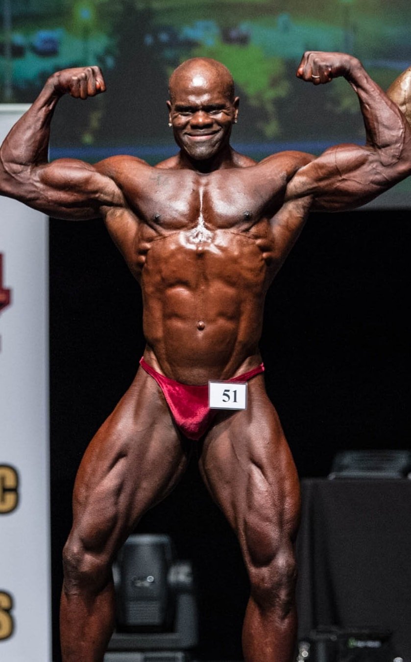 An older male bodybuilder with dark skin flexing his muscles during a competition, wearing a red posing suit with the number 51, on a stage with a dark background. Benny Mobley Public Speaker Emcee Podcast Coach Washington training on on one mentorsh