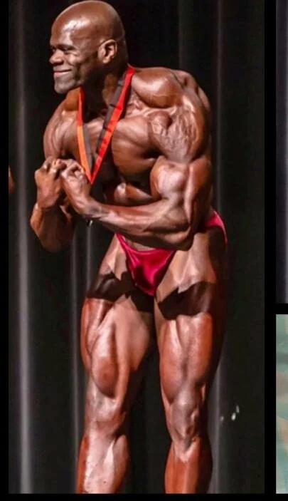 A male bodybuilder on stage in a red posing suit, flexing his arm muscles and wearing a medal around his neck. Benny Mobley Public Speaker Emcee Podcast Coach Washington training on on one mentorship