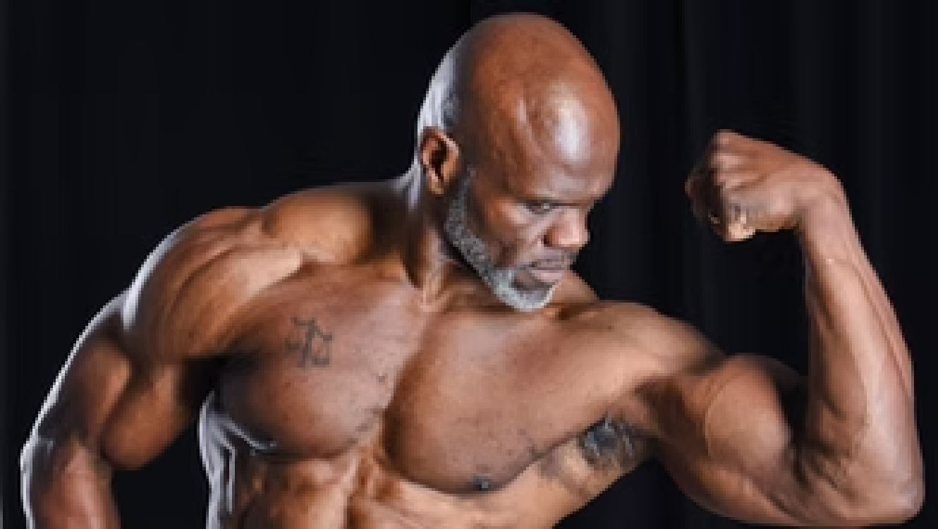 An older muscular man with a shaved head and gray beard flexing his bicep at a photography studio against a black background. Benny Mobley Public Speaker Emcee Podcast Coach Washington training on on one mentorship