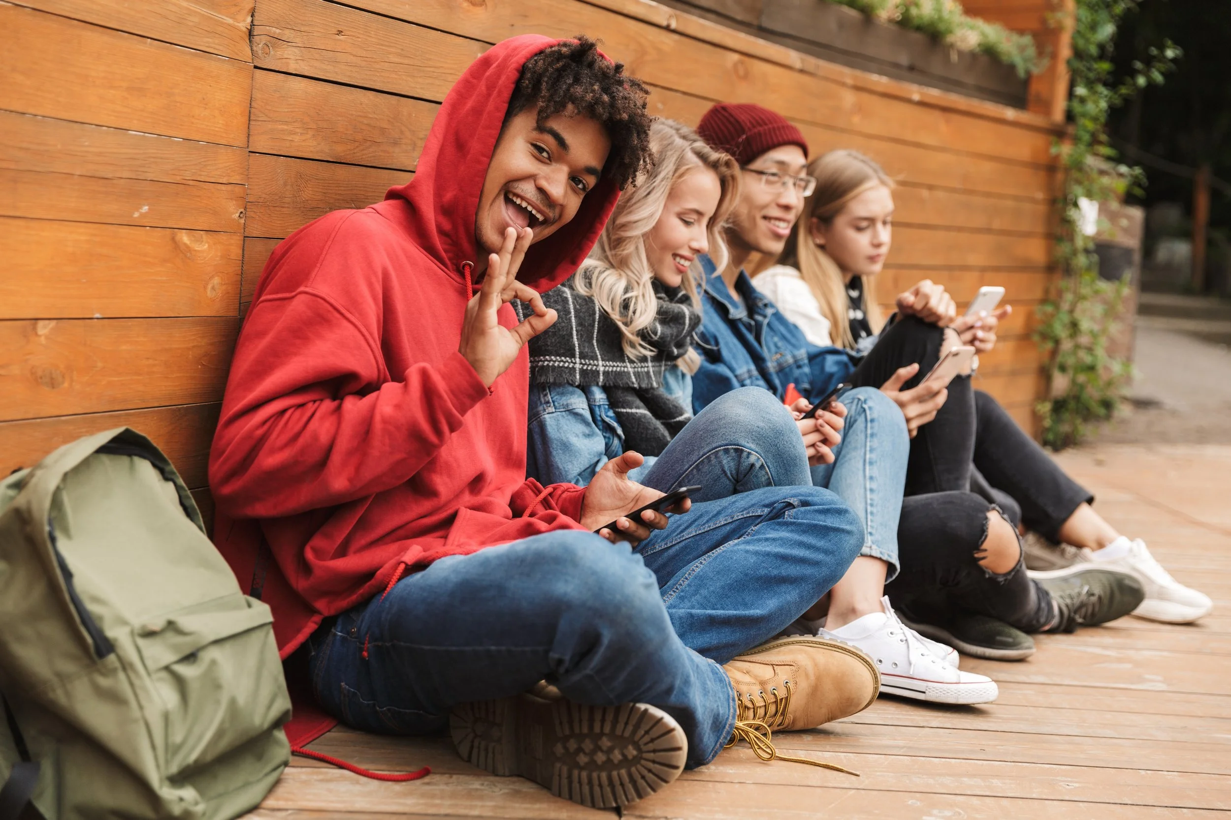 A group of five young friends sitting on a wooden deck outdoors, all looking at their smartphones and smiling. Benny Mobley Public Speaker Emcee Podcast Coach Washington training on on one mentorship