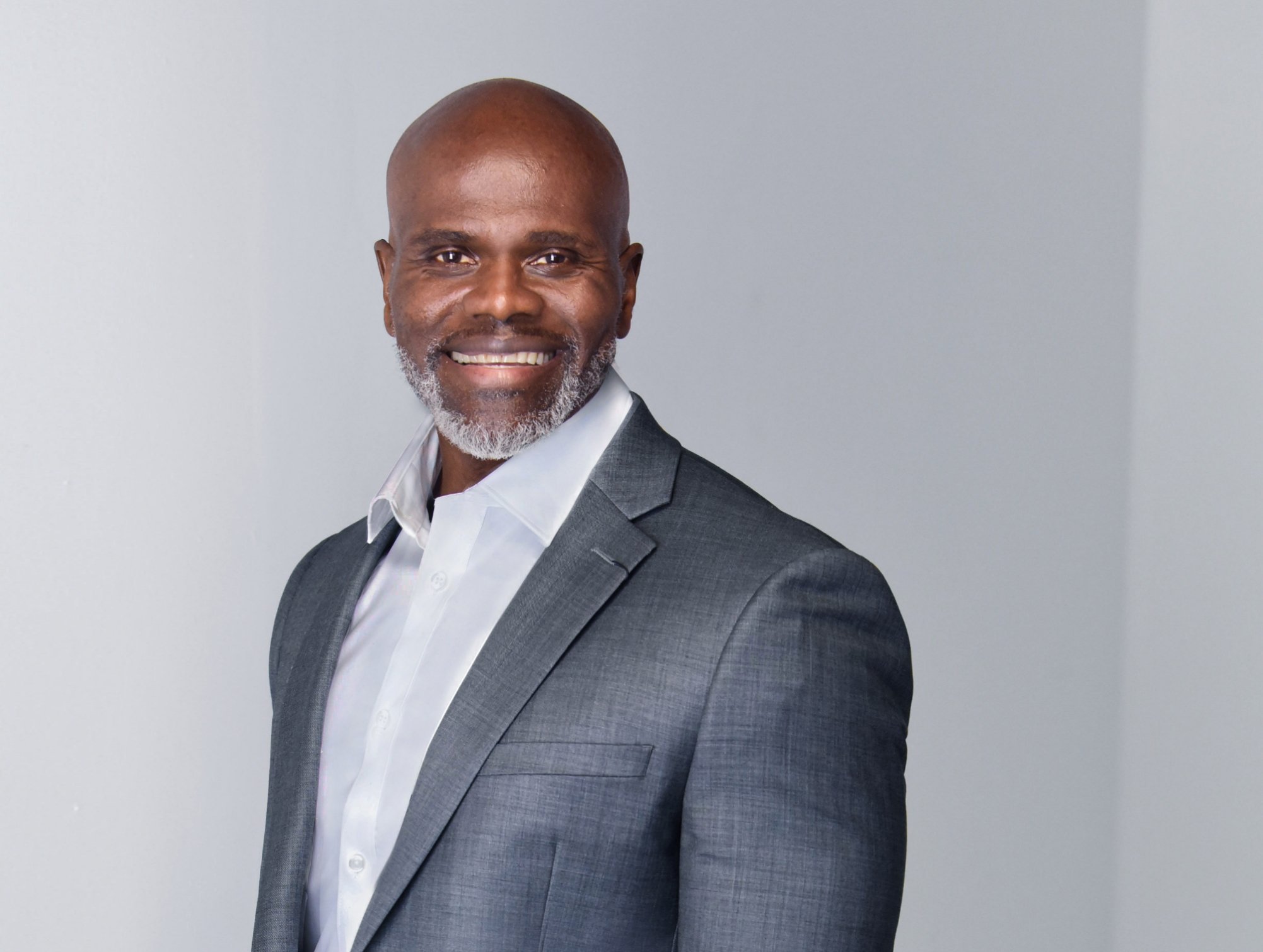 A smiling middle-aged man with a gray beard, wearing a gray suit and white shirt, standing against a plain light gray background. Benny Mobley Public Speaker Emcee Podcast Coach Washington training on on one mentorship