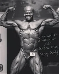 Black male bodybuilder flexing muscles on stage with a smile, wearing a competition number 51. Benny Mobley Public Speaker Emcee Podcast Coach Washington training on on one mentorship