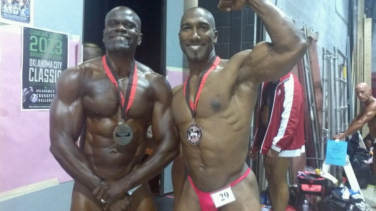 Two muscular men with medals around their necks posing together, one flexing his arm, in a gym or competition setting. Benny Mobley Public Speaker Emcee Podcast Coach Washington training on on one mentorship