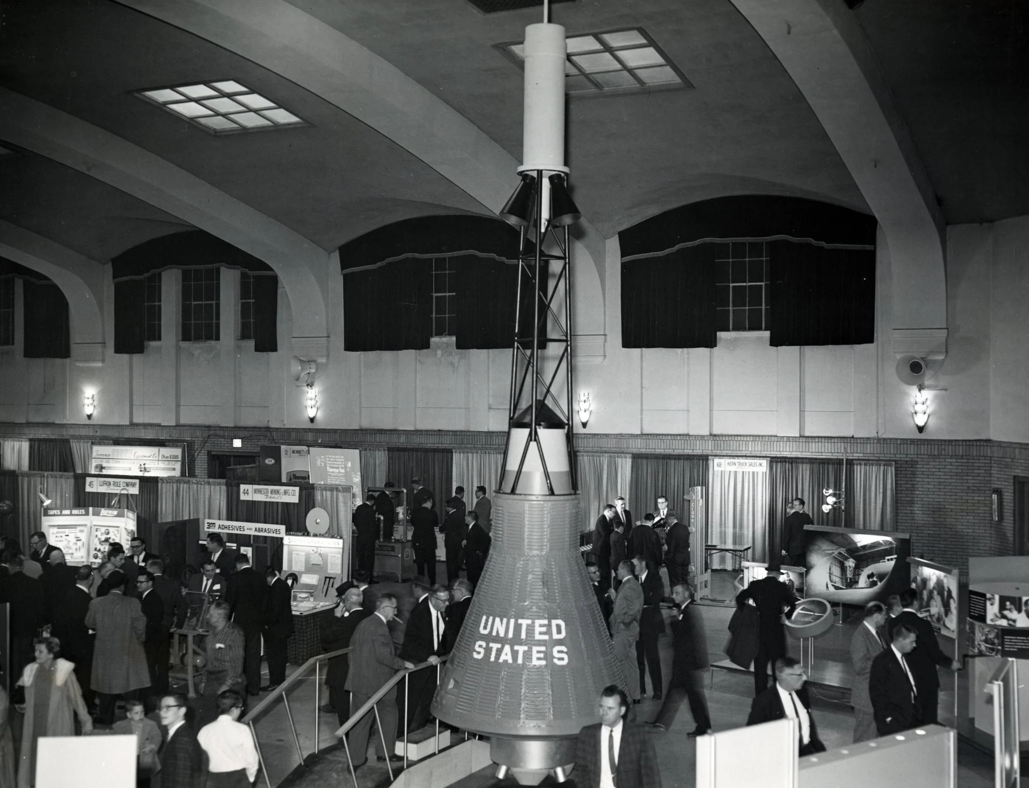 Black and white photo of a crowded exhibition hall with numerous people walking around and viewing various displays. A large model of a rocket labeled 'United States' stands prominently at the center of the hall. The hall has high ceilings and windows with dark curtains.