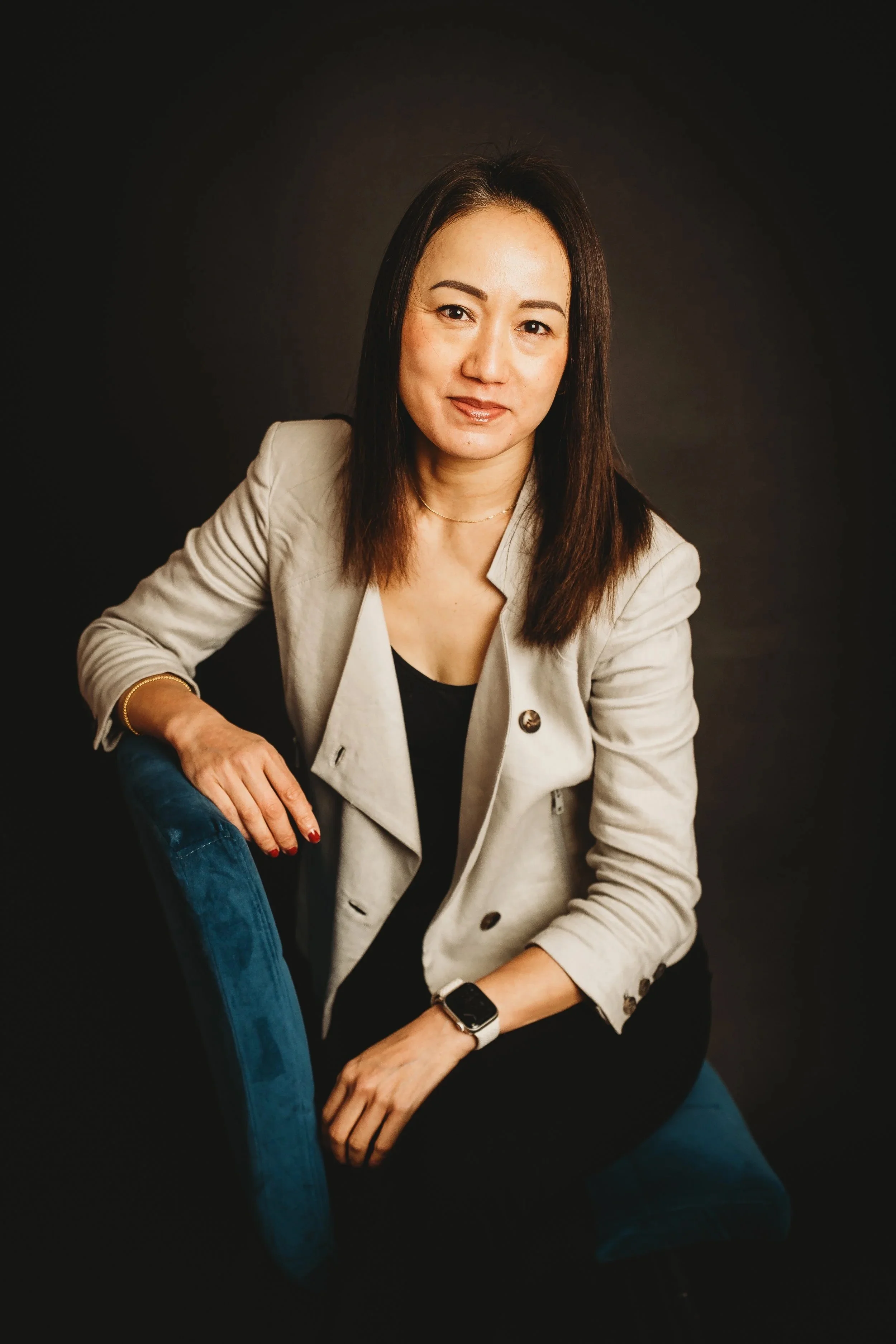 A woman with shoulder-length brown hair, wearing a beige blazer and black top, sitting on a blue chair against a dark background, looking at the camera.