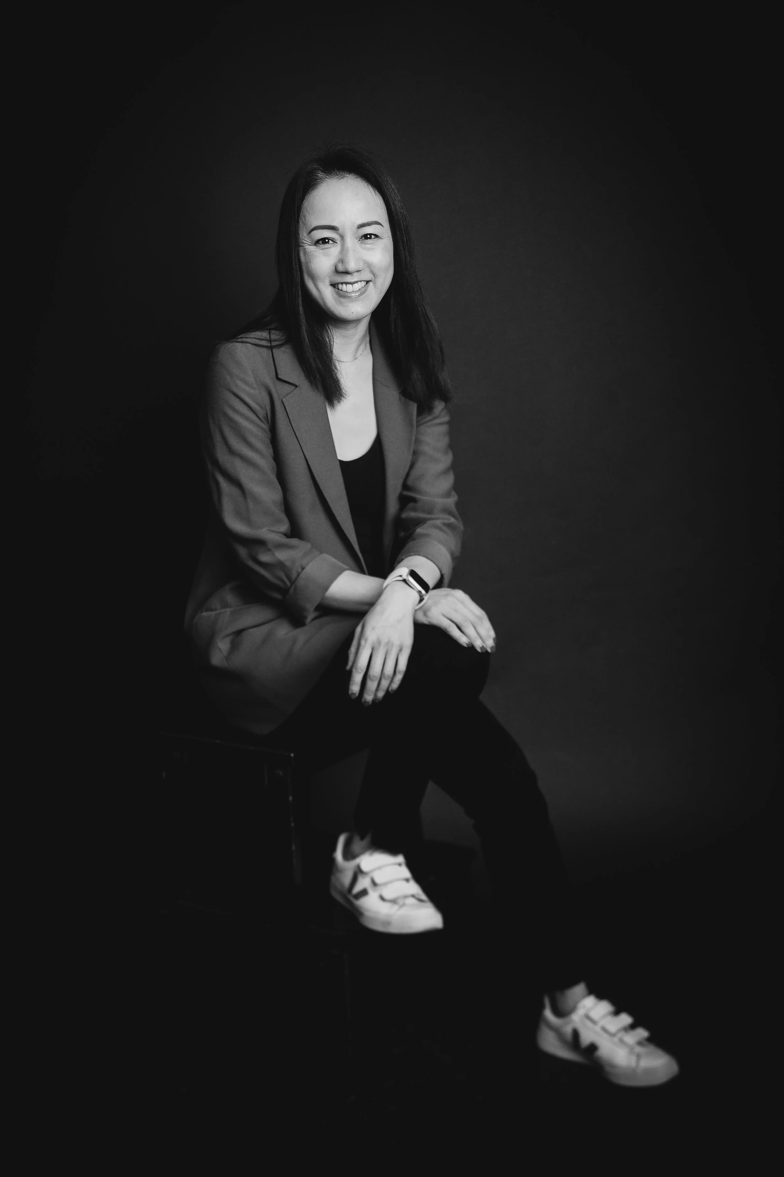 Black and white photo of a female psychotherapist smiling, seated on a chair a, dressed in a blazer, black top, and sneakers, with a smart/ casual appearance.