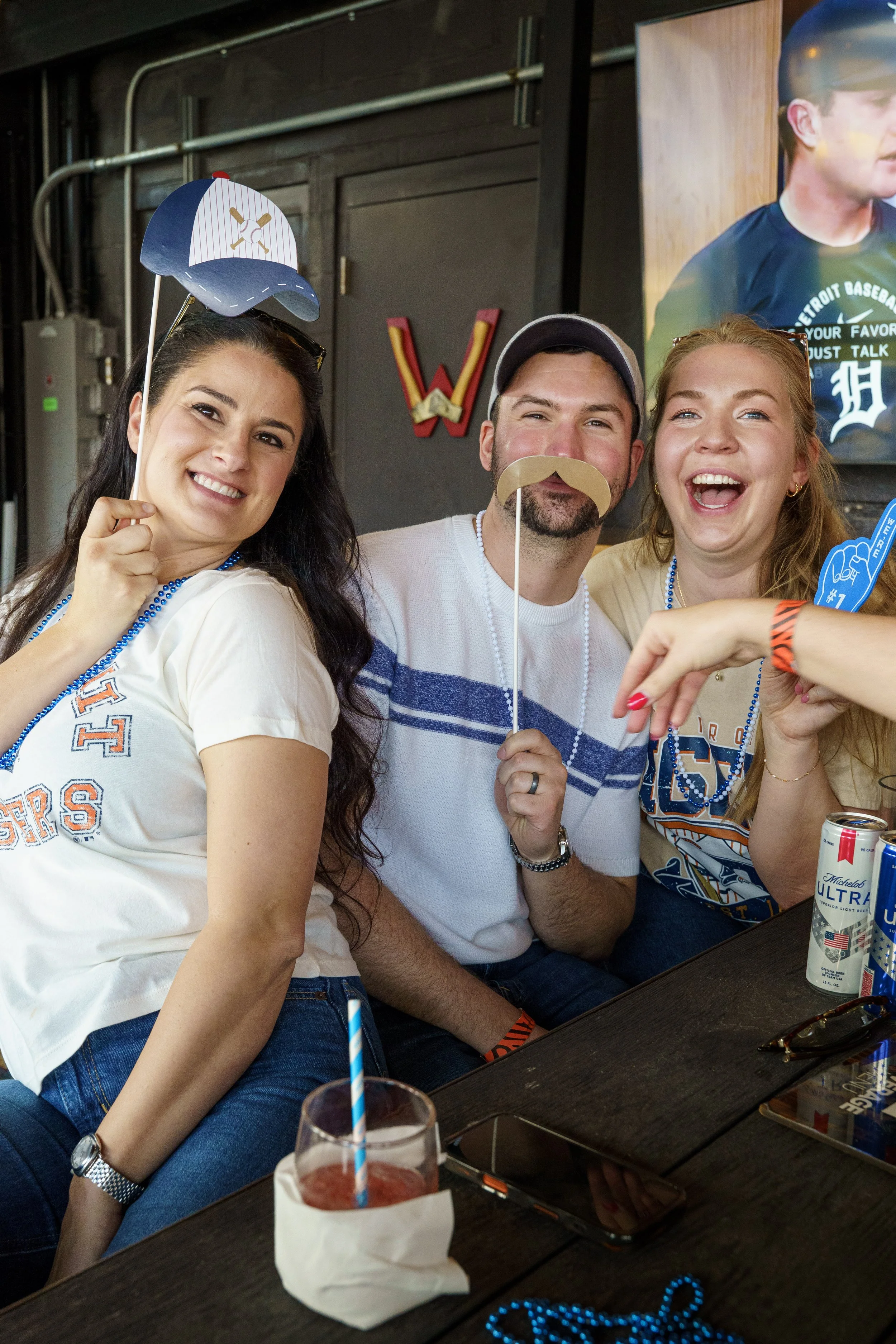 Three friends sitting at a table, smiling and holding fun photo props like a baseball cap and a fake mustache. The woman on the left is holding a paper baseball cap prop, the man in the middle has a paper mustache, and the woman on the right is laughing, holding a blue foam finger. Drinks and sunglasses are on the table. They are indoors, with a TV and a wall with a red letter "W" in the background.