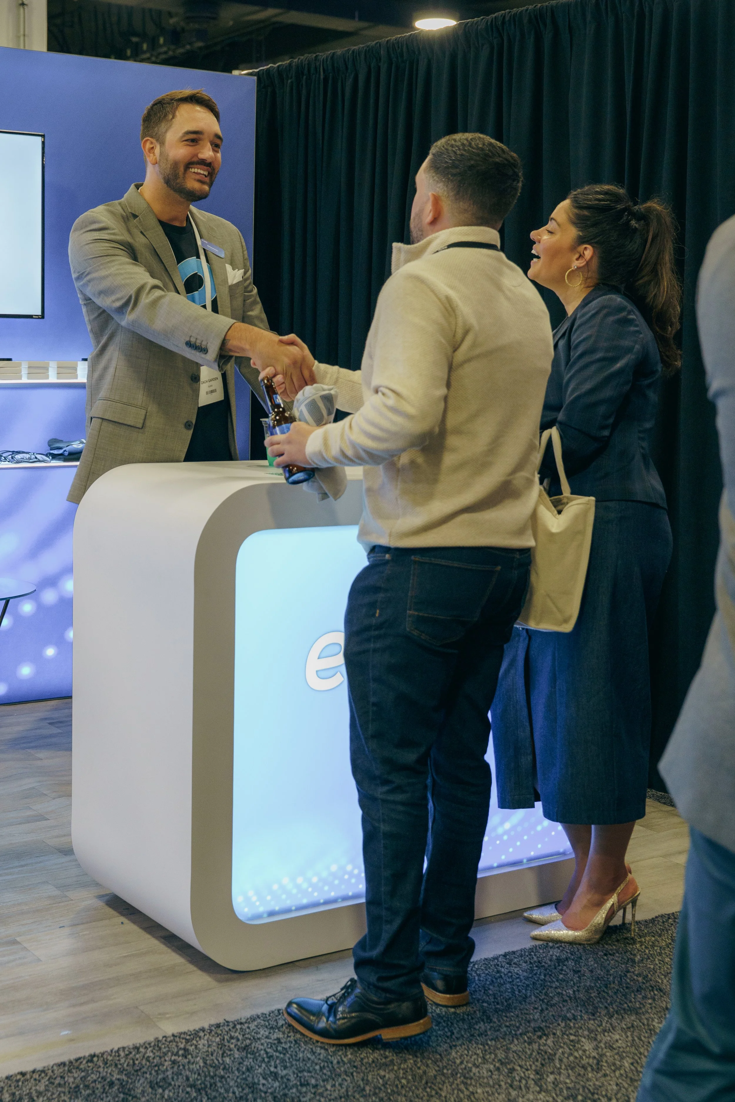 A man in a beige jacket and jeans shaking hands with a man in a gray suit at a booth with a blue background. A woman in a blue blazer and high heels stands next to them, smiling.