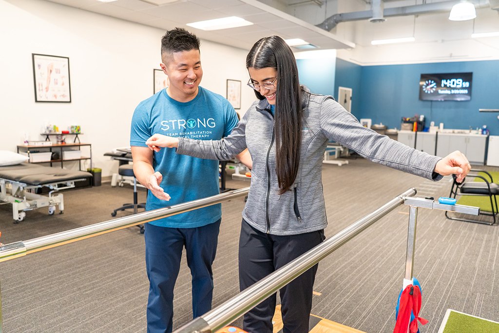 A woman in a gray jacket performs a balance exercise on parallel bars with assistance from a male therapist in a rehab center.