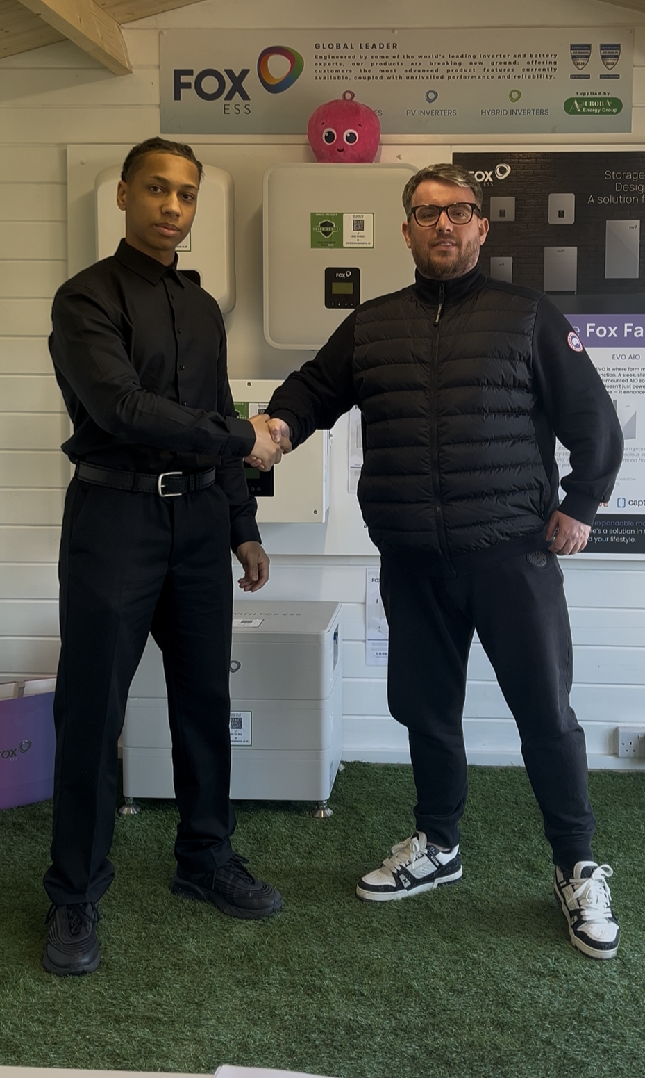 Two men shaking hands in front of a display of solar equipment, one young man dressed in black and the other in black athletic wear, inside a room with white walls and green carpet.
