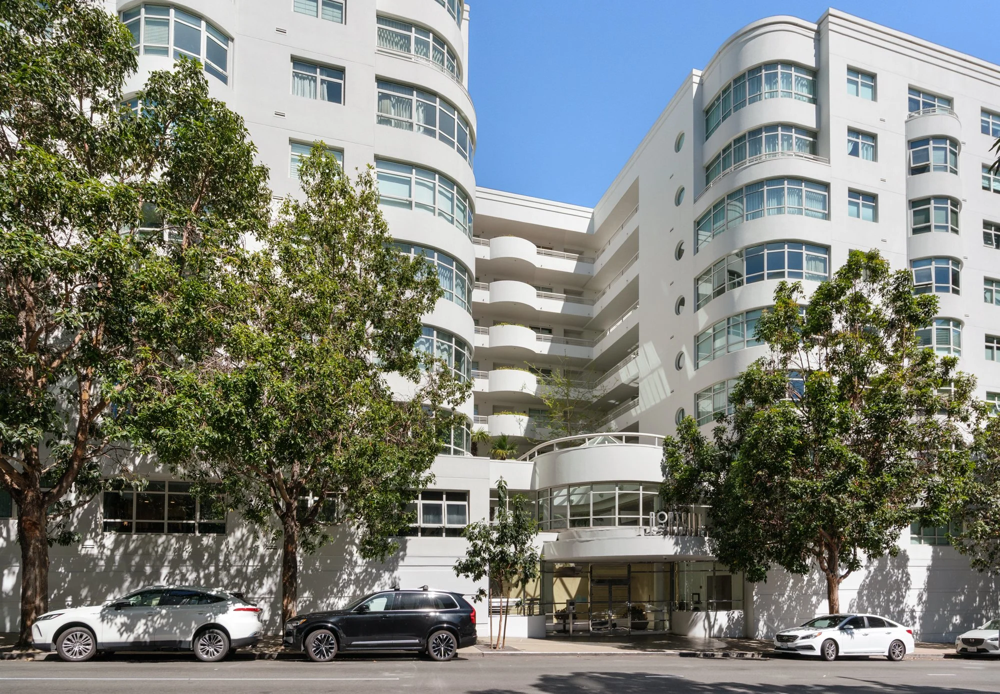White multi-story residential building with rounded balconies and large windows, trees lining the sidewalk, parked cars in front, and a clear blue sky above.