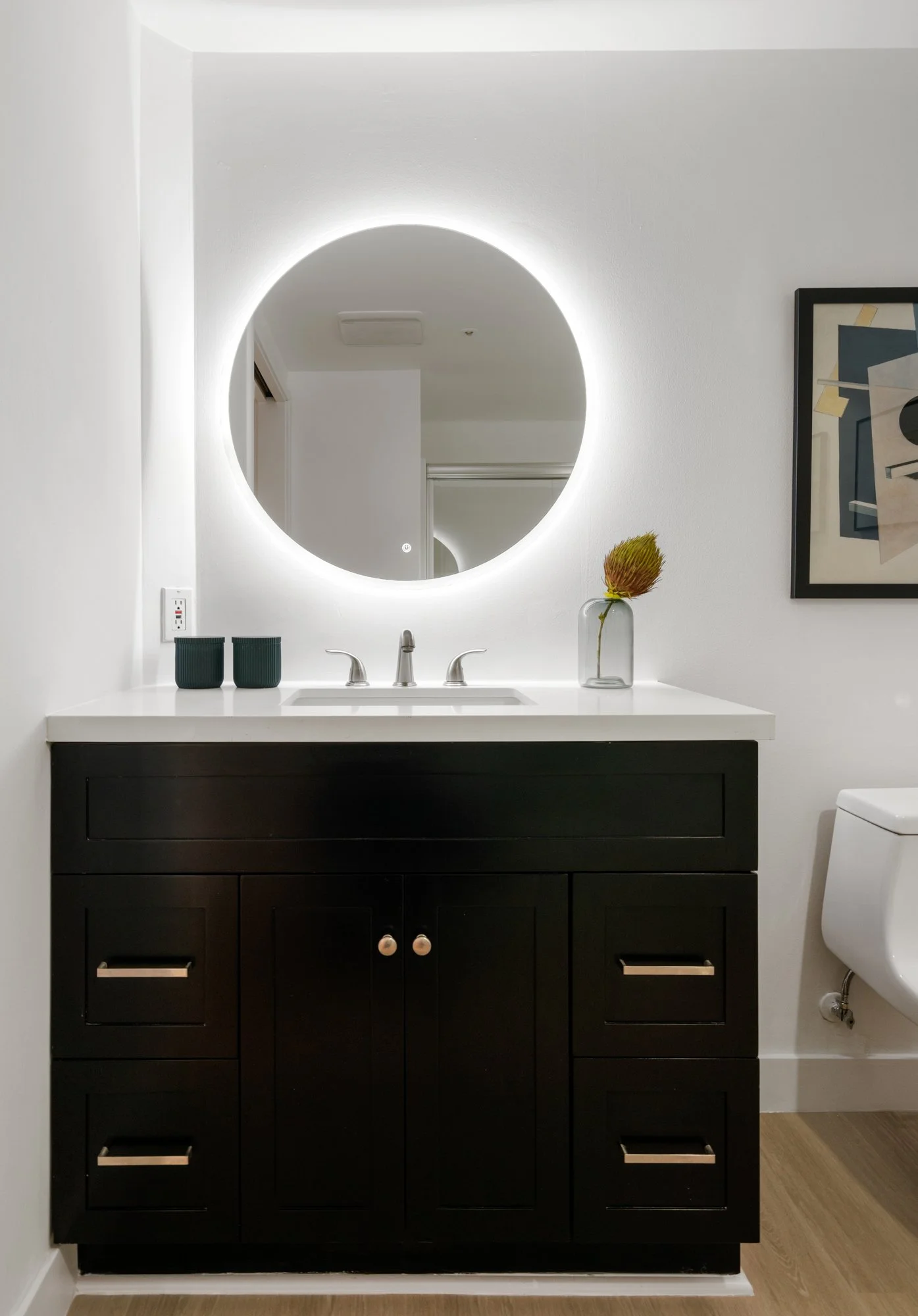Modern bathroom vanity with black cabinets, white countertop, and a round backlit mirror. A small vase with a flower and two cups are on the countertop, with a framed abstract art piece on the wall to the right.