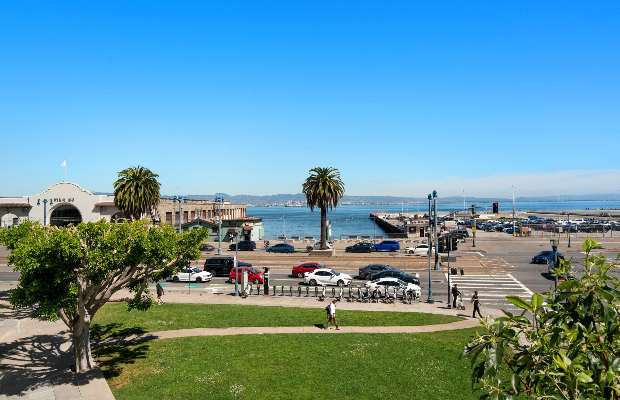 A view of a waterfront area with palm trees, cars, pedestrians, and a pier with water and a distant city skyline under a clear blue sky.