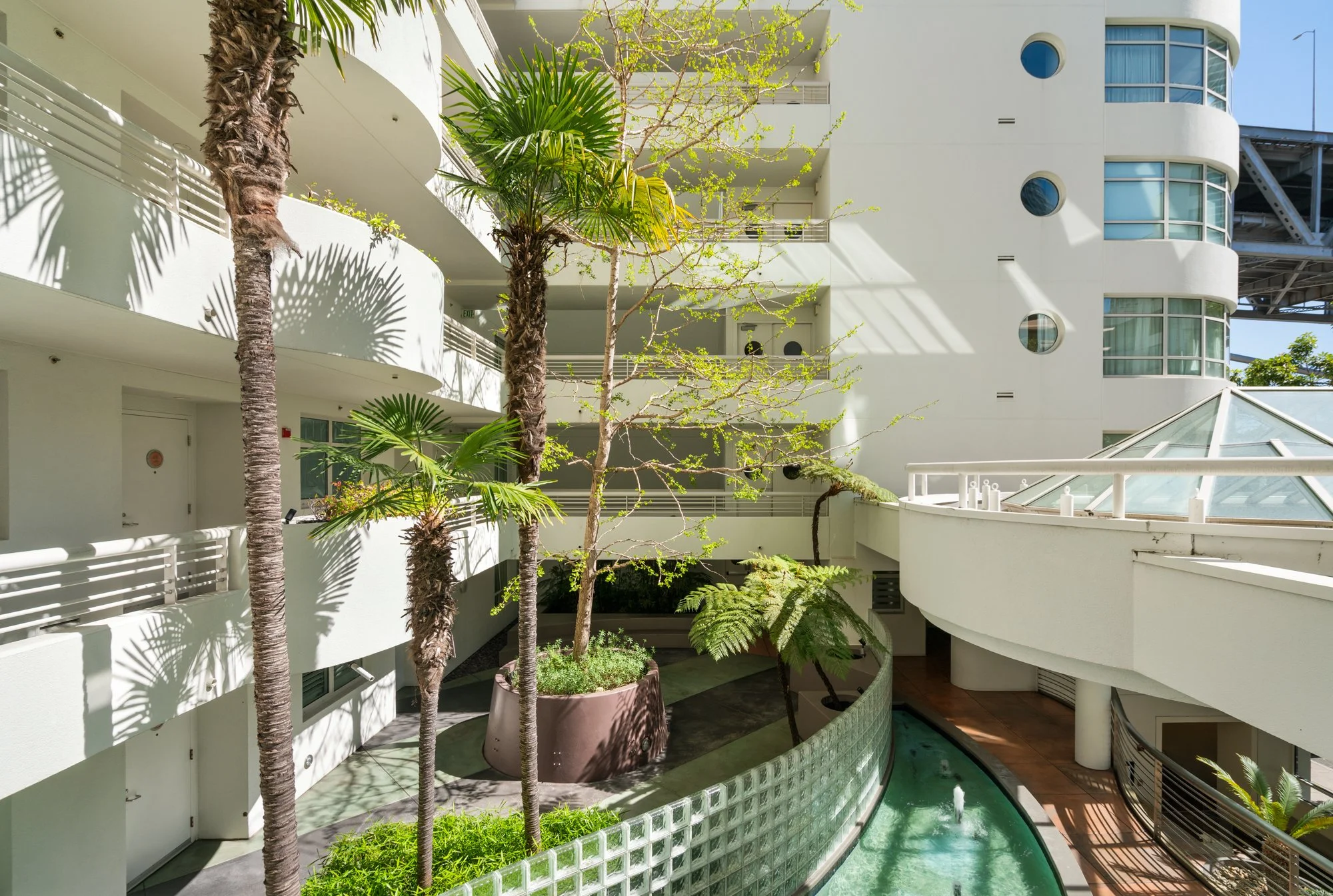Interior courtyard with white buildings, green palm and fern trees, balcony railings, and a curved pool with a glass block wall, sunlight creating shadows.