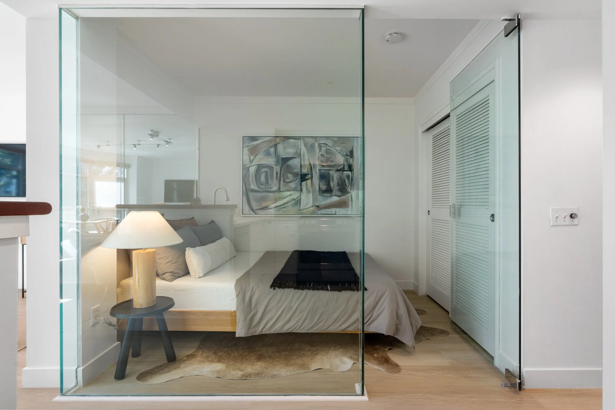 Modern bedroom behind a glass wall with bed, nightstand, lamp, and artwork