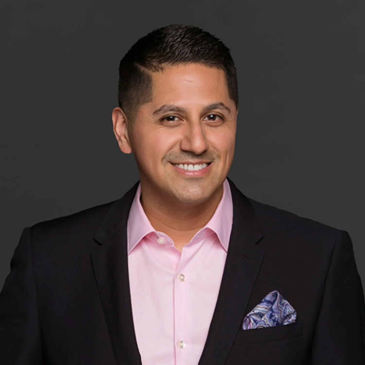 A smiling man in a black suit jacket and pink dress shirt with a blue patterned pocket square, against a dark grey background.