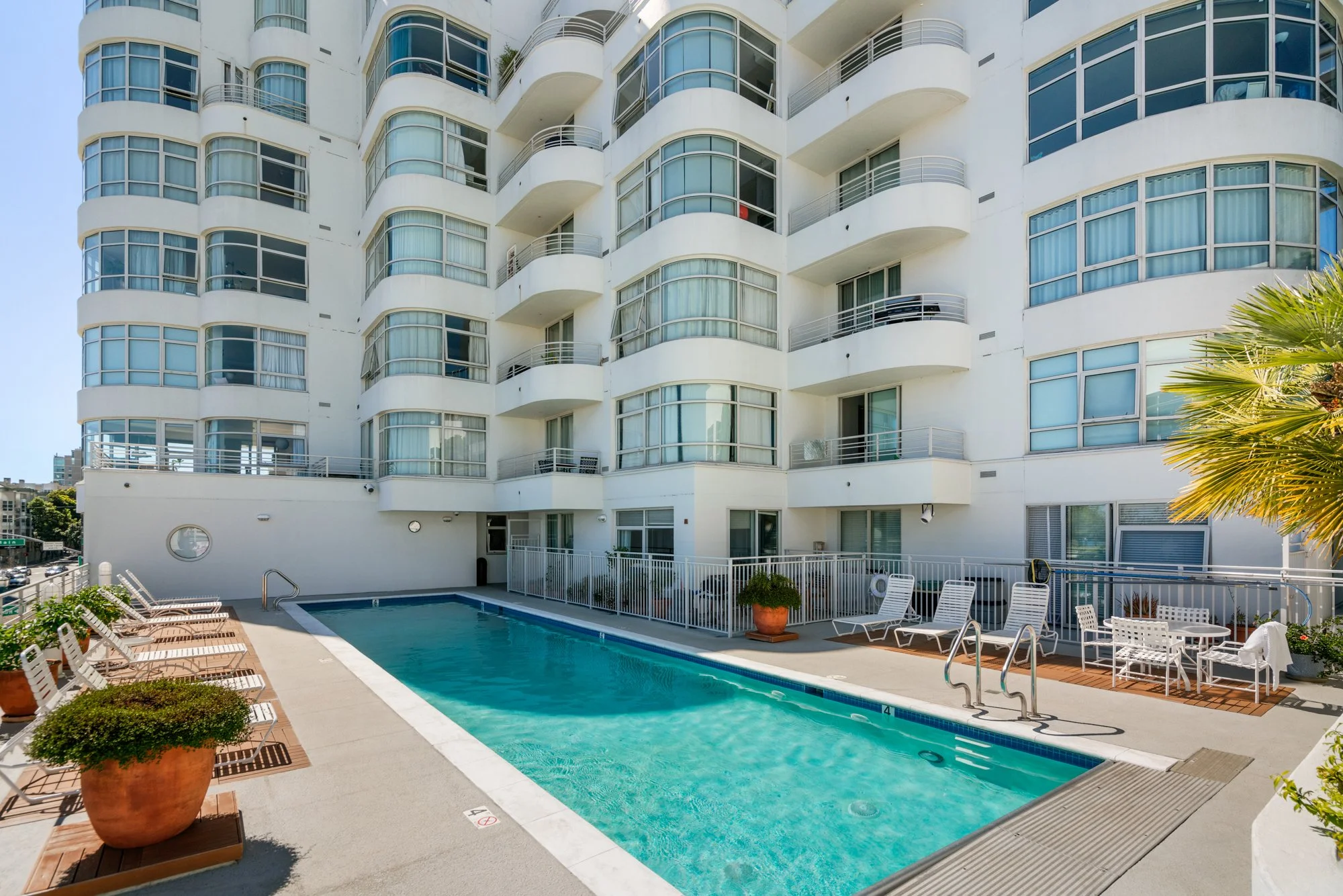 Outdoor swimming pool area in front of a modern, white, multi-story building with curved balconies, outdoor lounge chairs, and potted plants.