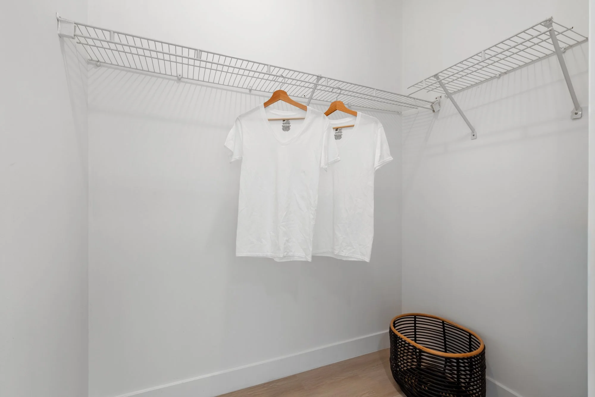 Empty walk-in closet with a white wire shelf, two white t-shirts on hangers, and a black woven laundry basket.