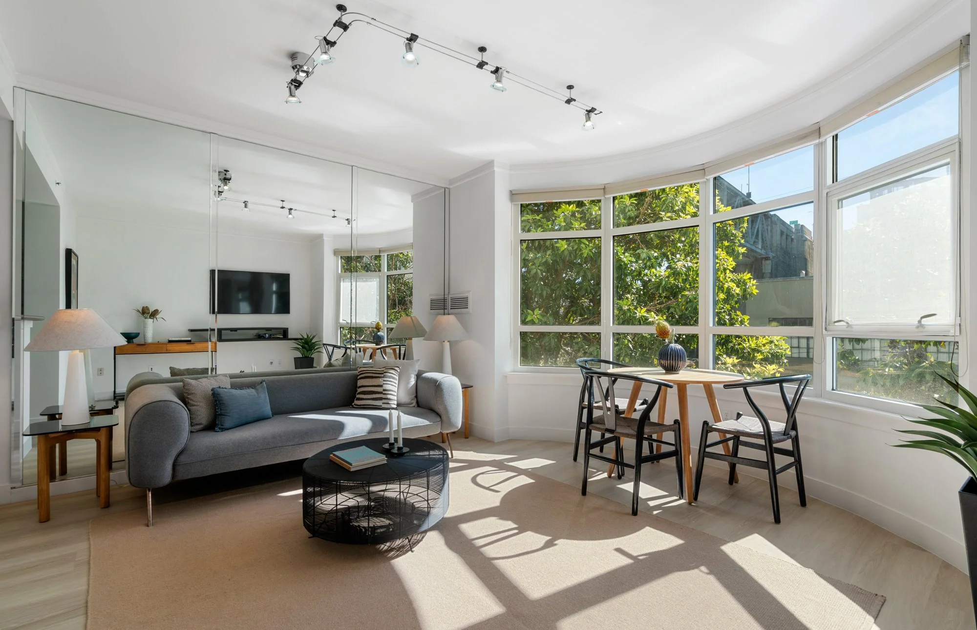 Bright living room with large bay windows, gray sofa with pillows, and a black coffee table with books and candles. There is a round wooden dining table with four black chairs, and sunlight creates shadows on the beige rug.
