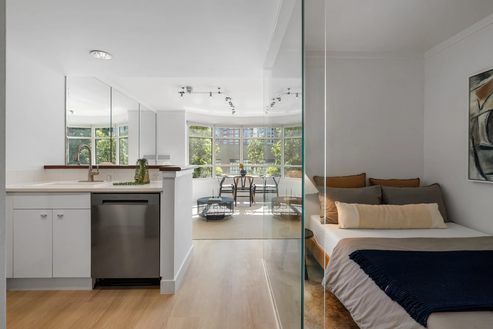 Open-plan living space with a kitchen, dining area, and a bedroom separated by a glass partition. The kitchen has white cabinets, a stainless steel dishwasher, and a countertop with a plant. The dining area features a table and chairs near large windows with greenery outside. The bedroom contains a bed with multiple pillows and wall art.